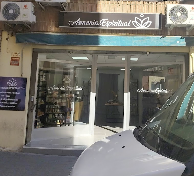 Fachada de tienda llamada 'Armonía Espiritual' con ventanas de vidrio y escritorios internos visibles, señal de servicios espirituales y productos como aceites naturales y velas aromáticas, ubicada en una calle.