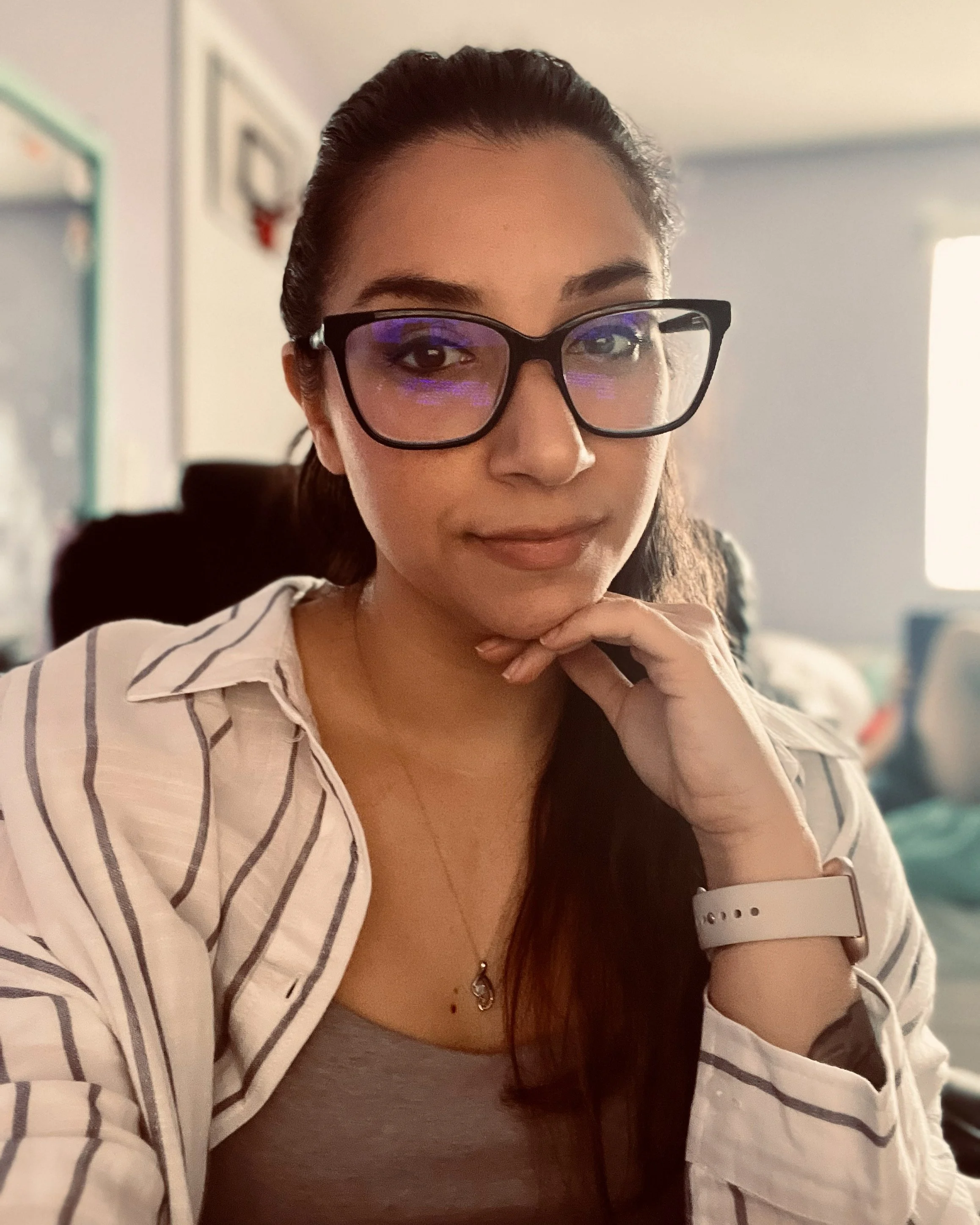 A woman wearing glasses with purple reflection, a striped white and gray shirt over a gray top, and a watch on her left wrist, sitting indoors with her hand resting on her chin.