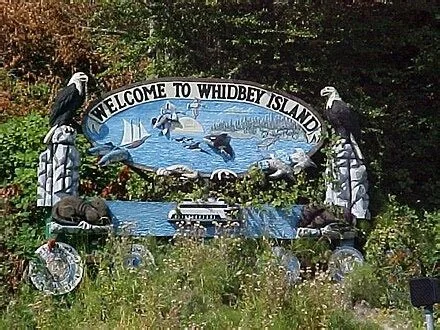 Welcome sign for Whidbey Island with wildlife and water scenes, surrounded by rocks, plants, and physical decorations.