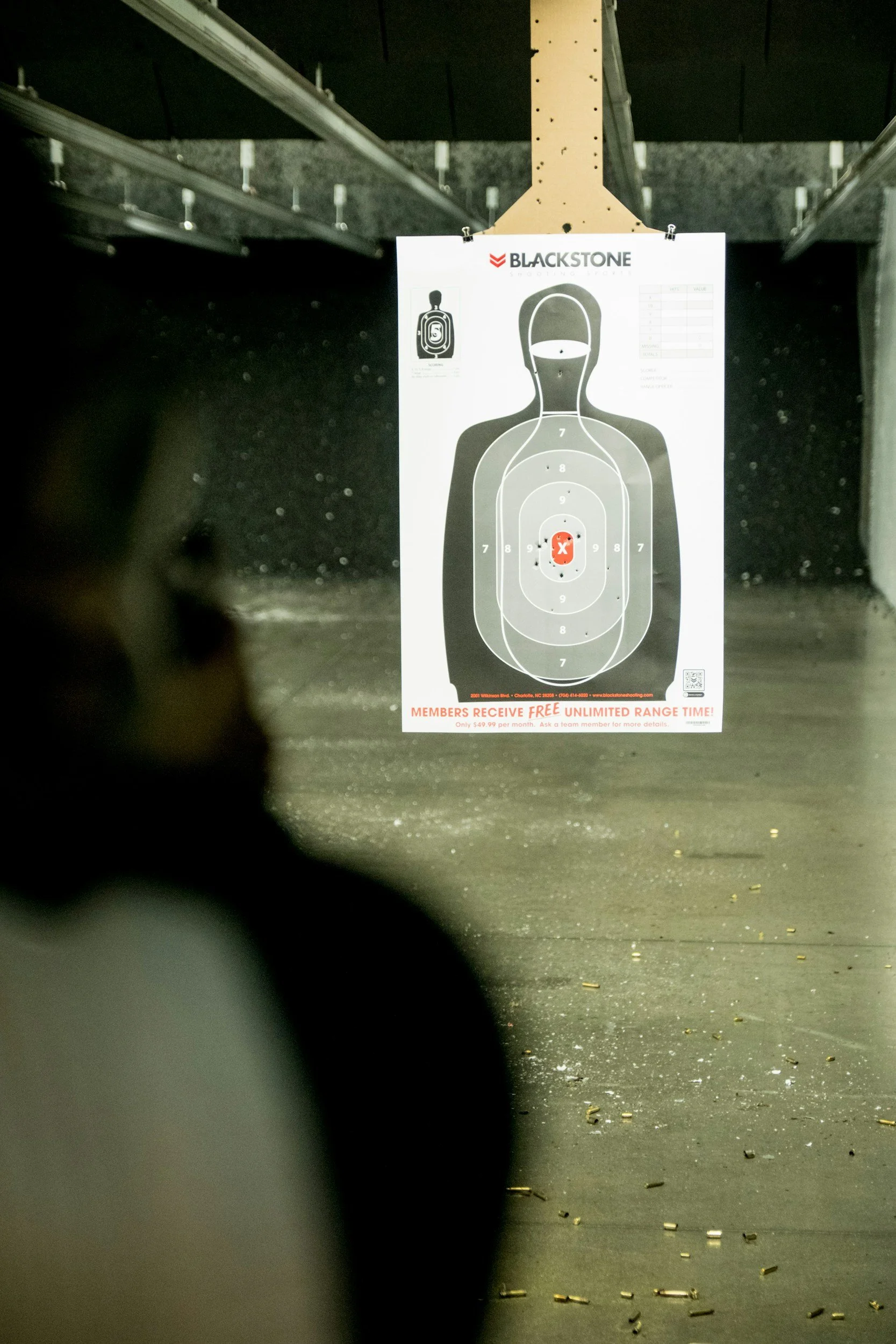 Target shooting range with a paper target hanging, showing multiple bullet holes, and a person's blurred silhouette in the foreground.
