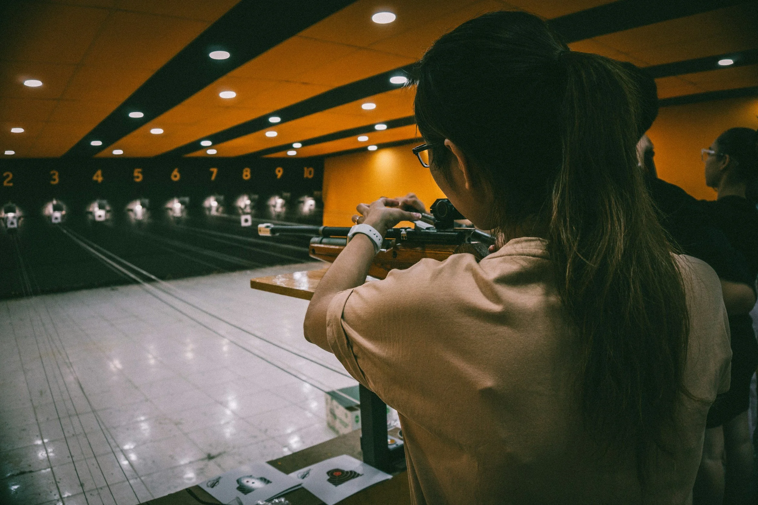 A woman at an indoor shooting range aiming a rifle towards target lanes numbered 2 to 10.