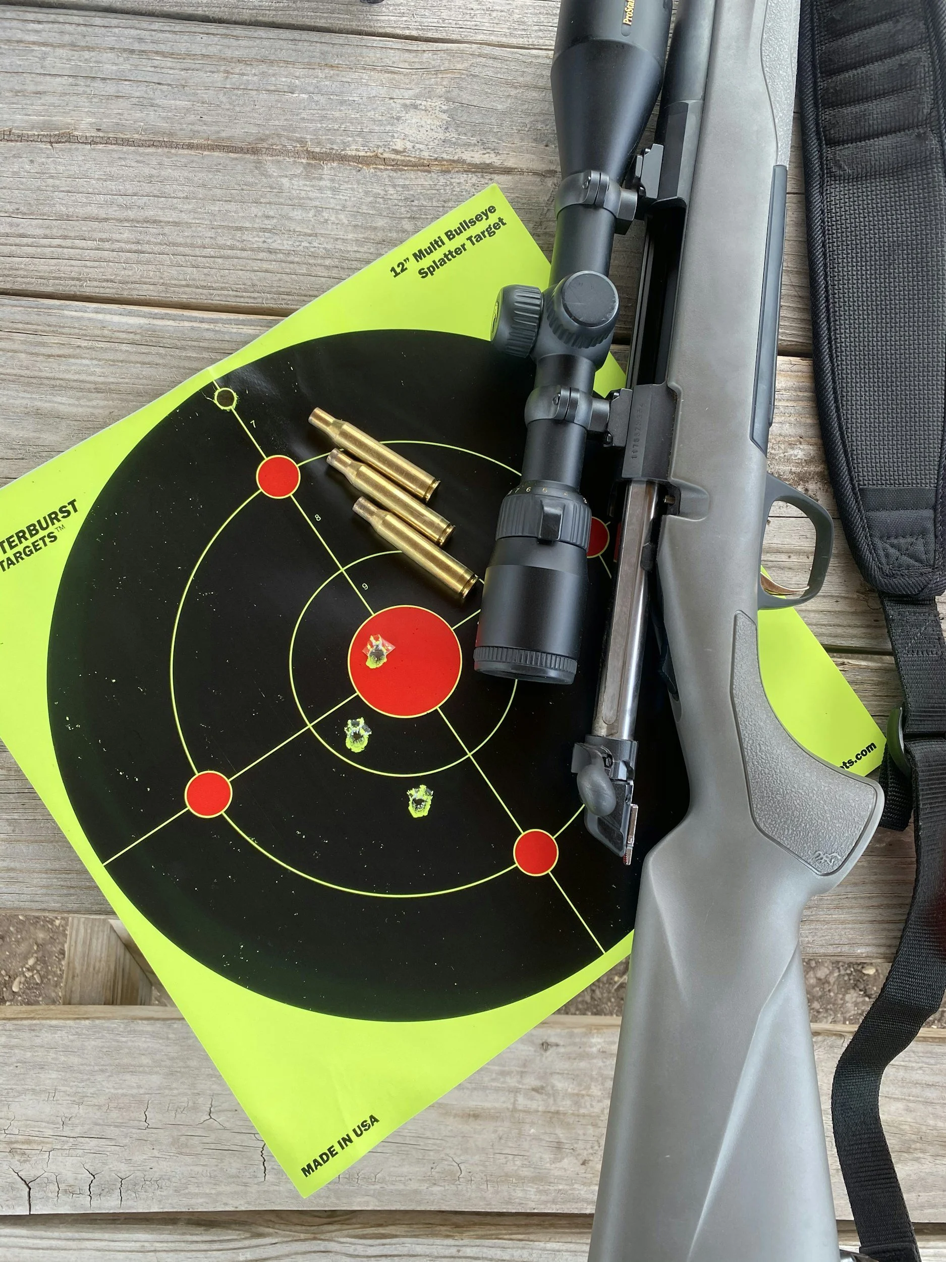 A firearm with a scope resting on a yellow shooting target with bullet holes, placed on a wooden surface.