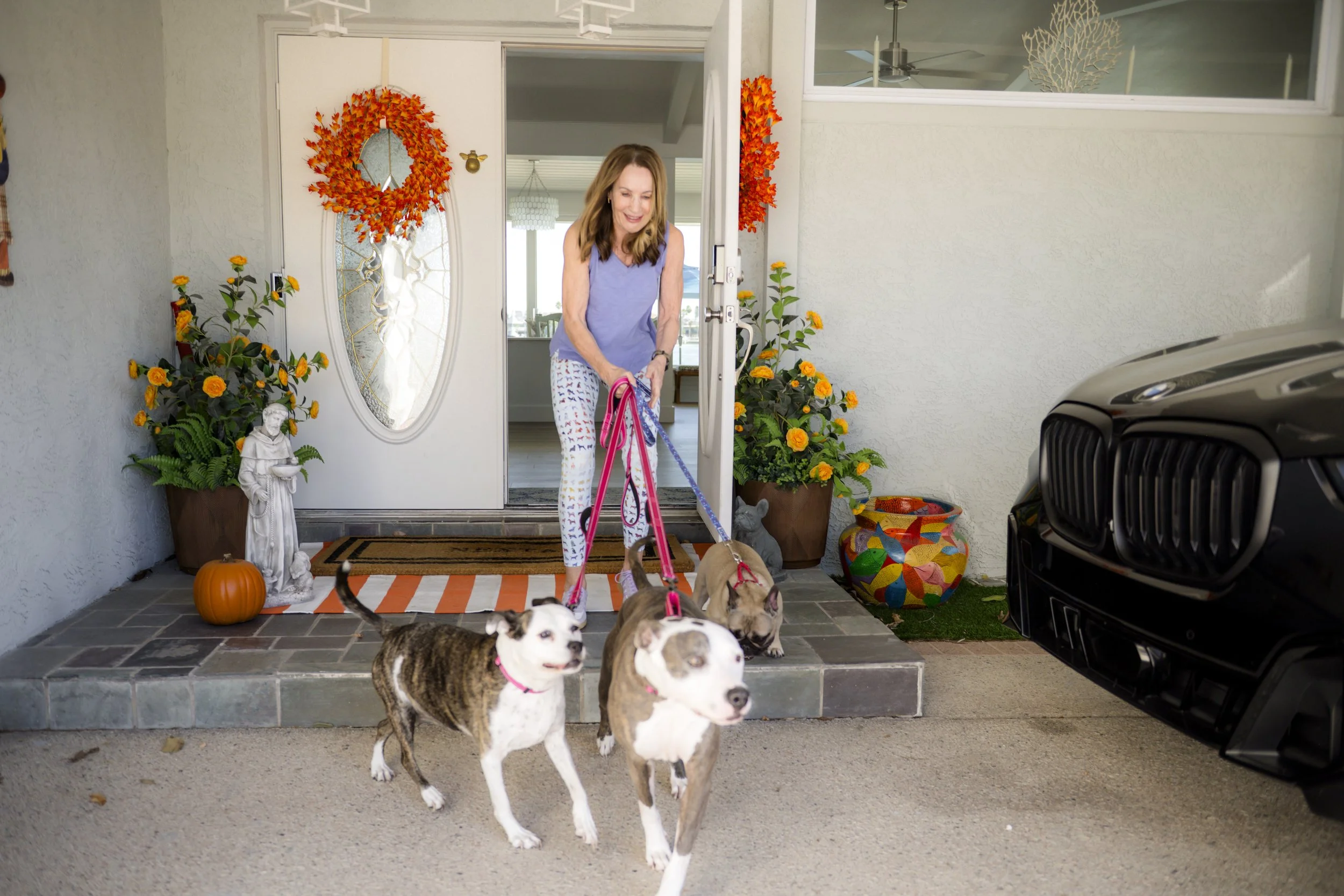 A woman is arriving home with three dogs, standing on the front porch beside potted plants, a pumpkin, and a colorful decorated pot, with a black car partially visible to the right.