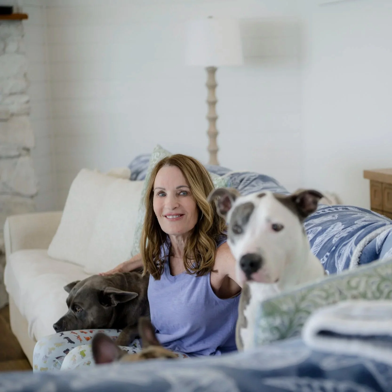 A woman with light brown hair sitting on a couch with two dogs, one black and one white with black spots, in a cozy living room.