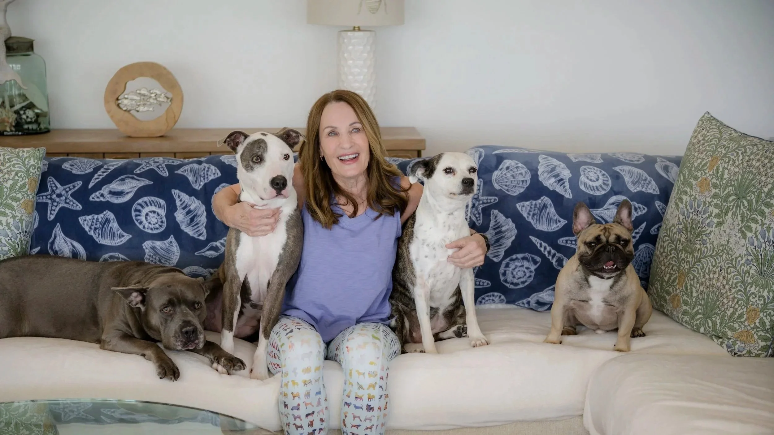 A woman sitting on a cream-colored couch with five dogs, smiling and hugging two of the dogs, with seashell-patterned cushions and a blue throw on the couch, in a living room with a wooden side table, decorative items, and a lamp behind her.