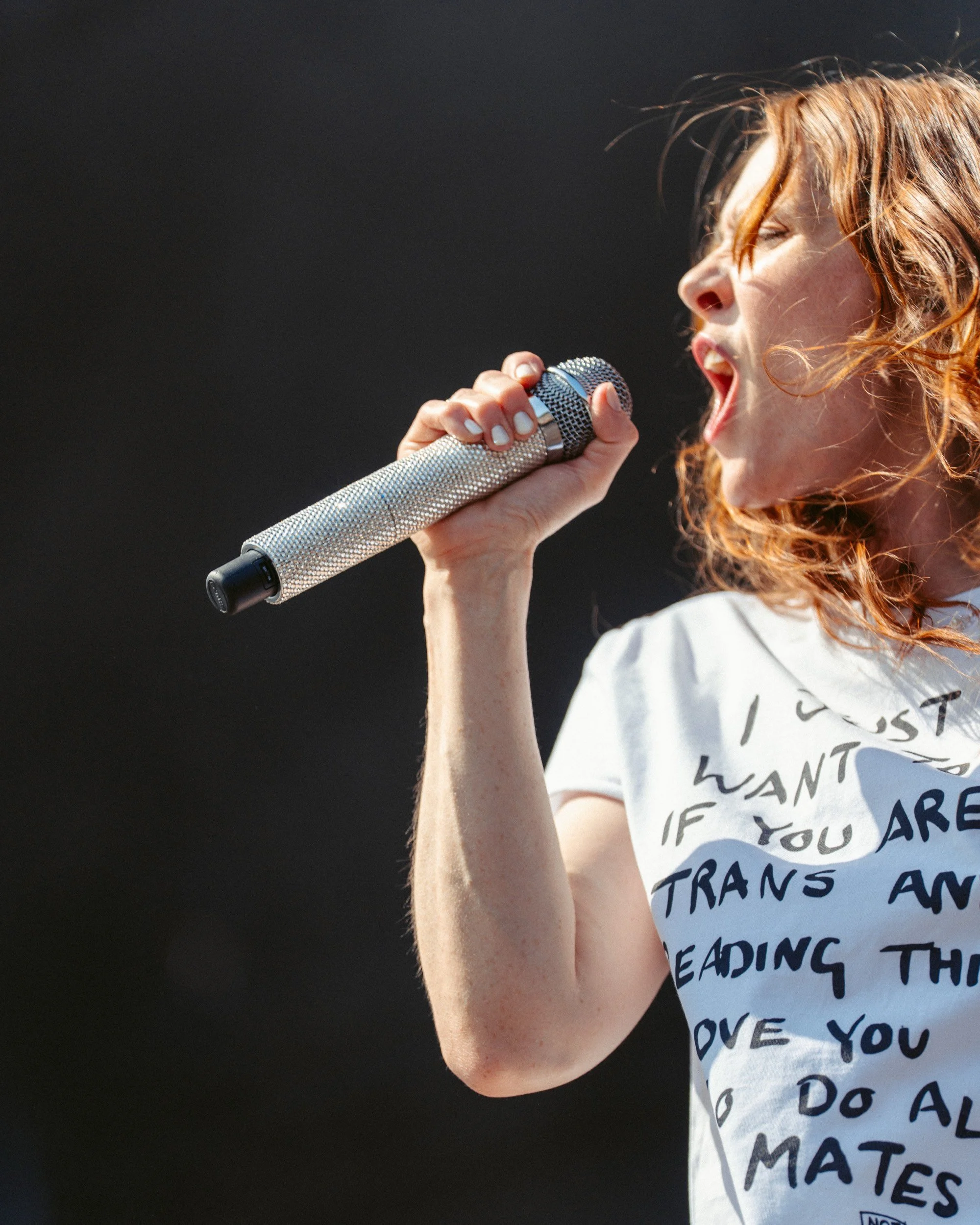 Junge Frau singt mit einem silbernen Mikrofon auf einer Bühne, trägt ein weißes T-Shirt mit schwarzem Text, rote Haare, Sonnenschein, dunkler Hintergrund.