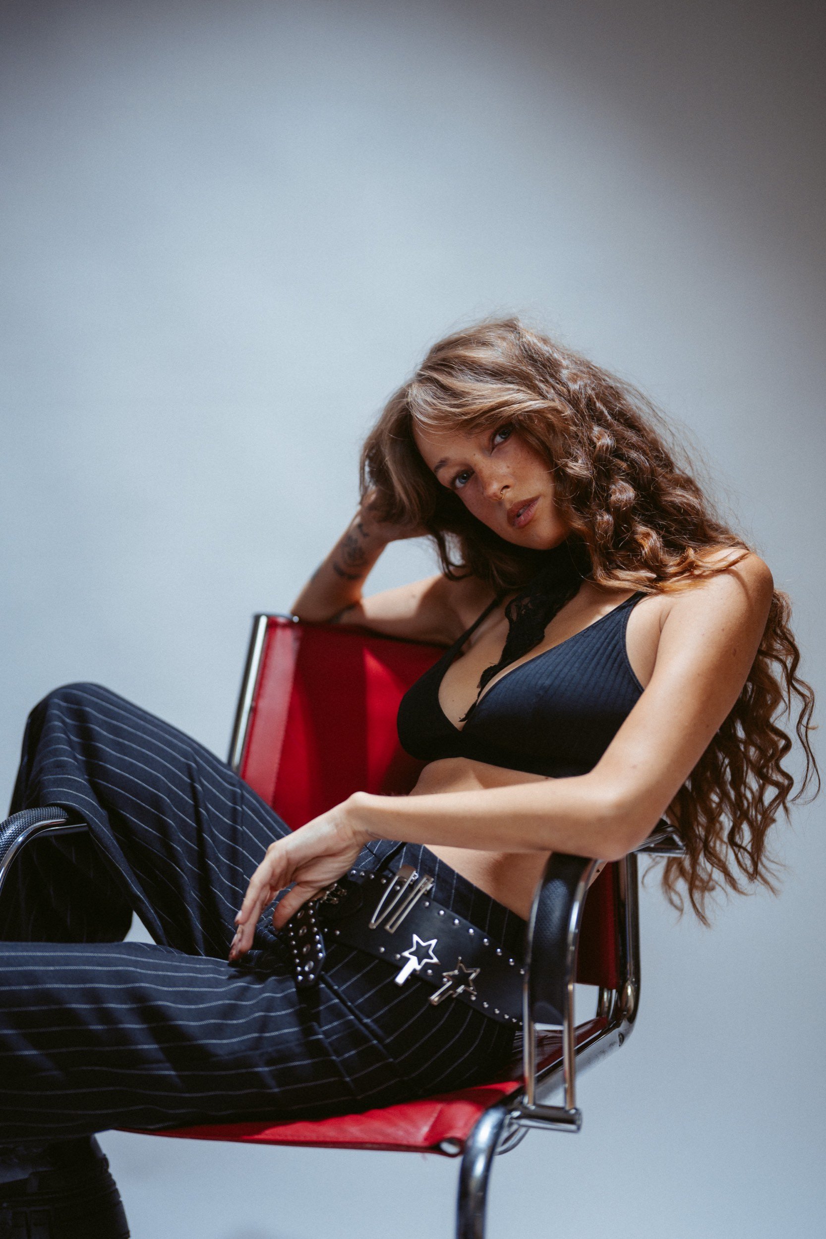 Junge Frau mit langen, lockigen Haaren sitzt entspannt in einem roten Stuhl, trägt ein schwarzes Top und schwarze, gestreifte Hosen mit einem schwarzen Gürtel mit Sternen- und Blitze-Details, vor grauem Hintergrund.