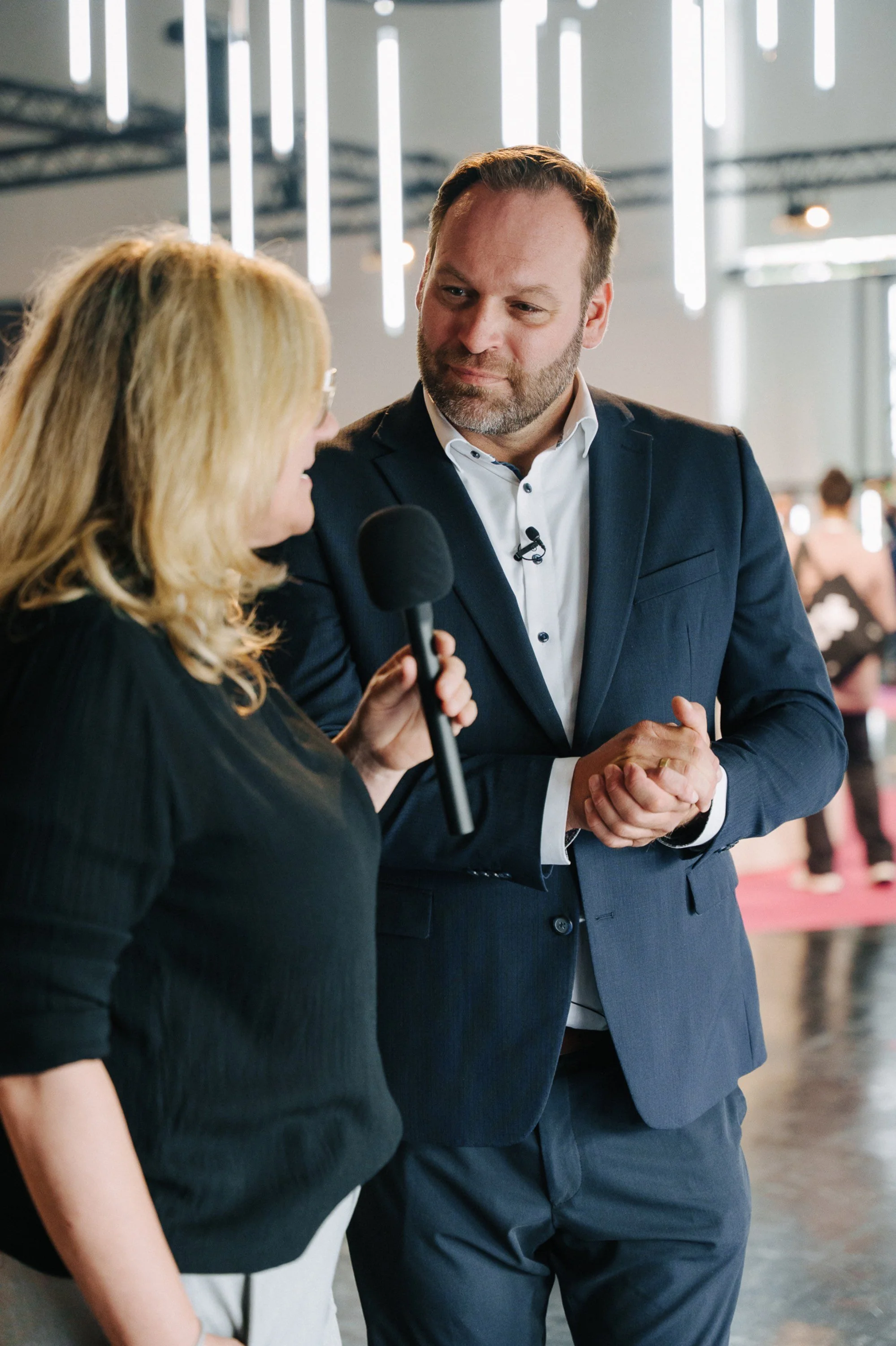 Ein Mann im Anzug spricht mit einer Frau, die einen Mikrofon hält, bei einer Veranstaltung im Innenraum.