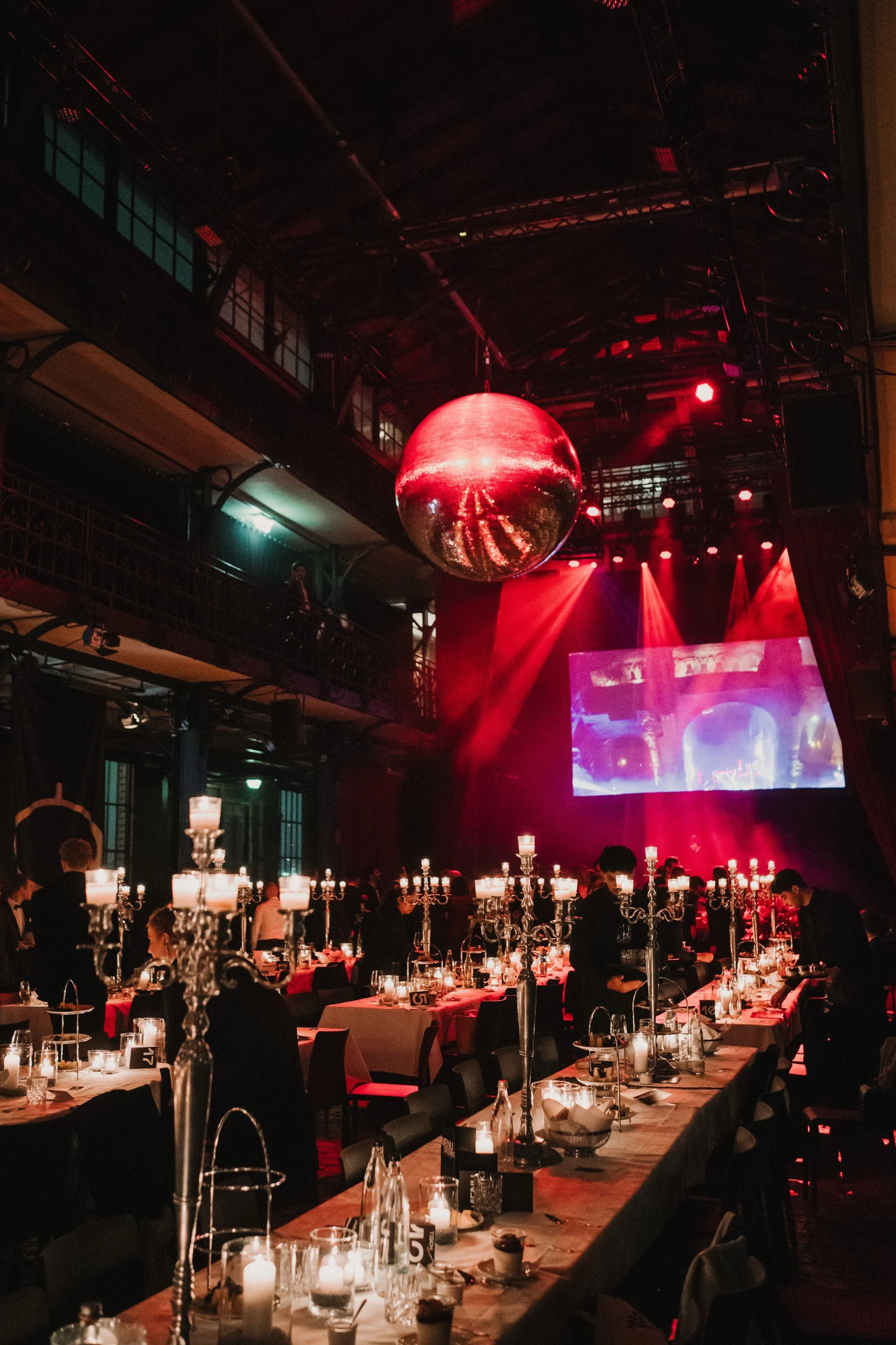 Eleganter Veranstaltungsraum mit rotem Beleuchtung, gespiegelt-kugel an Decke, lange Tafel mit Kerzen und Gäste, große Leinwand im Hintergrund.