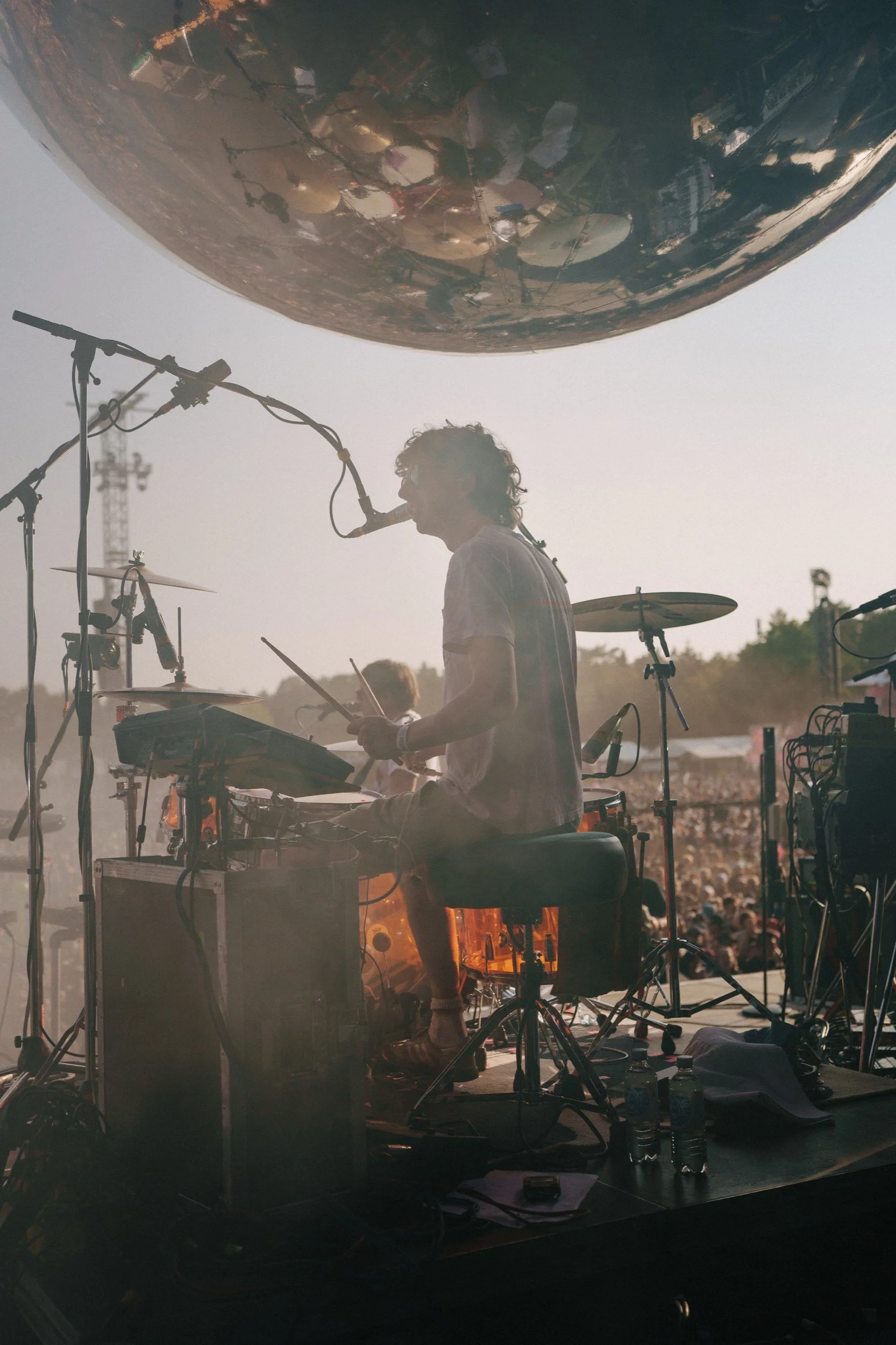 Musiker mit Trommelset und Mikrofon auf einer Bühne während eines Konzerts, im Hintergrund ein großes Publikum, Natur im Freien bei Sonnenuntergang.