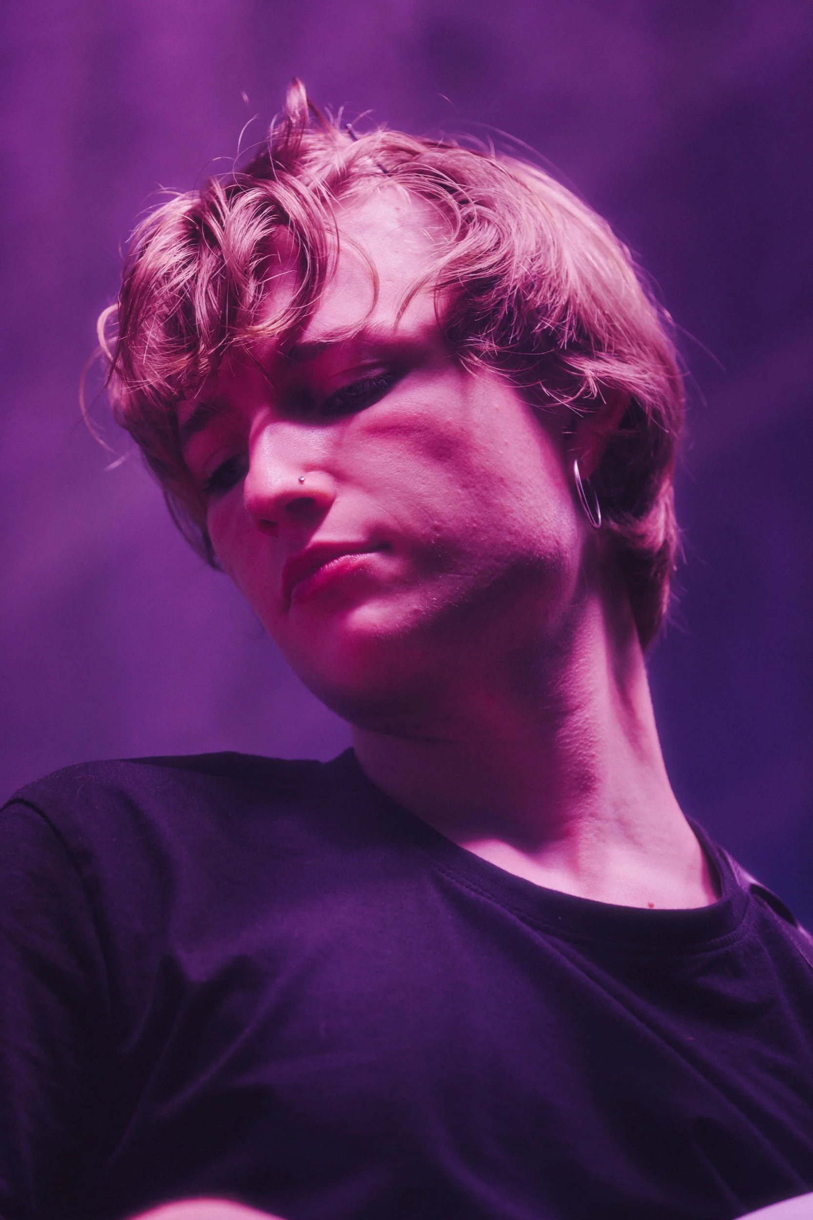 Nahaufnahme eines jungen Mannes mit lockigen, blonden Haaren, der in einem schwarzen T-Shirt under knalligem violettem Licht steht.