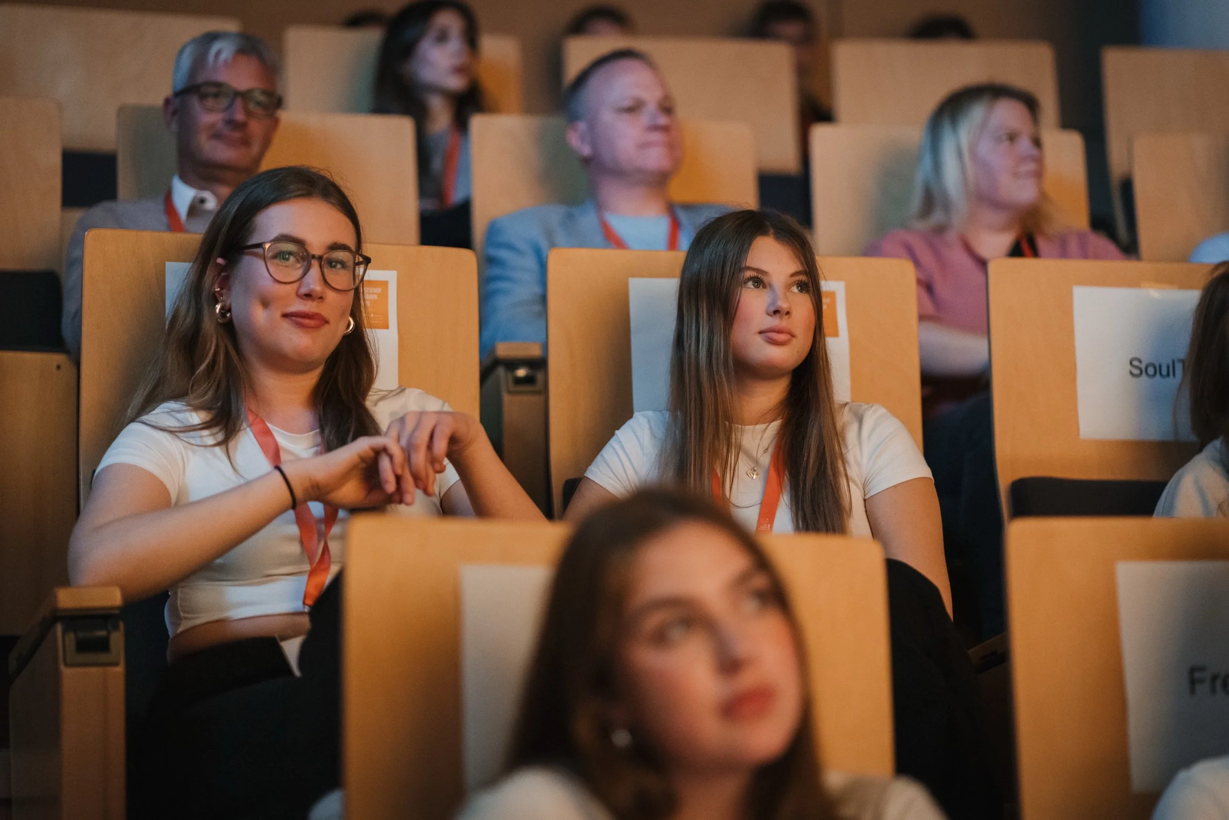 Menschen sitzen in einem Auditorium bei einer Konferenz oder Veranstaltung, einige schauen aufmerksam, einige nehmen Notizen oder haben nachdenkliche Ausdrücke.