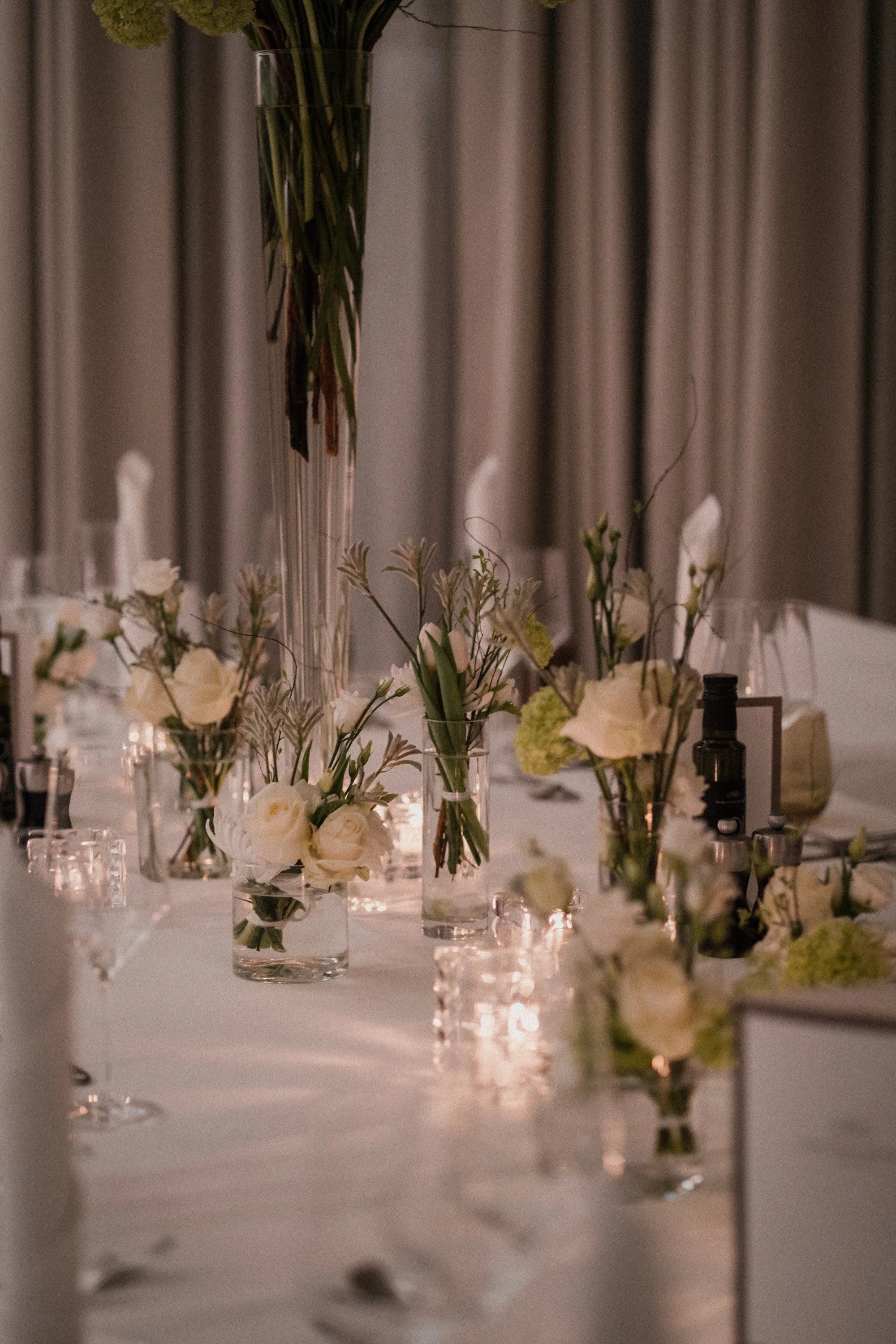 Ein eleganter Tisch mit weißen Blumen in verschiedenen Glasvasen, Teelichtern und dekorativen Objekten, vorbereitet für eine Feier in einem festlichen Raum.