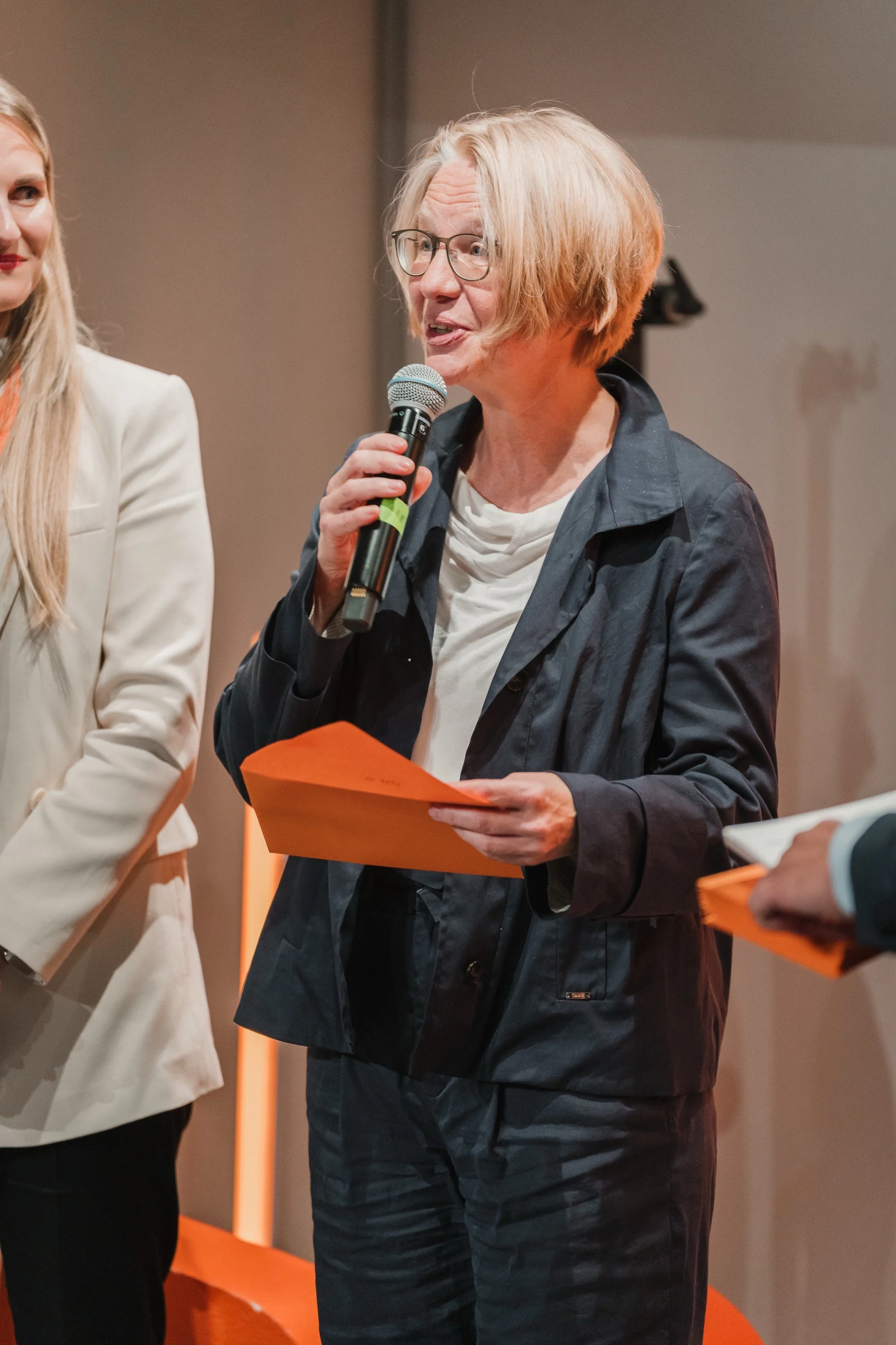 Eine ältere Frau mit kurz blondem Haar, Brille und dunkler Jacke spricht in ein Mikrofon bei einer Veranstaltung.
