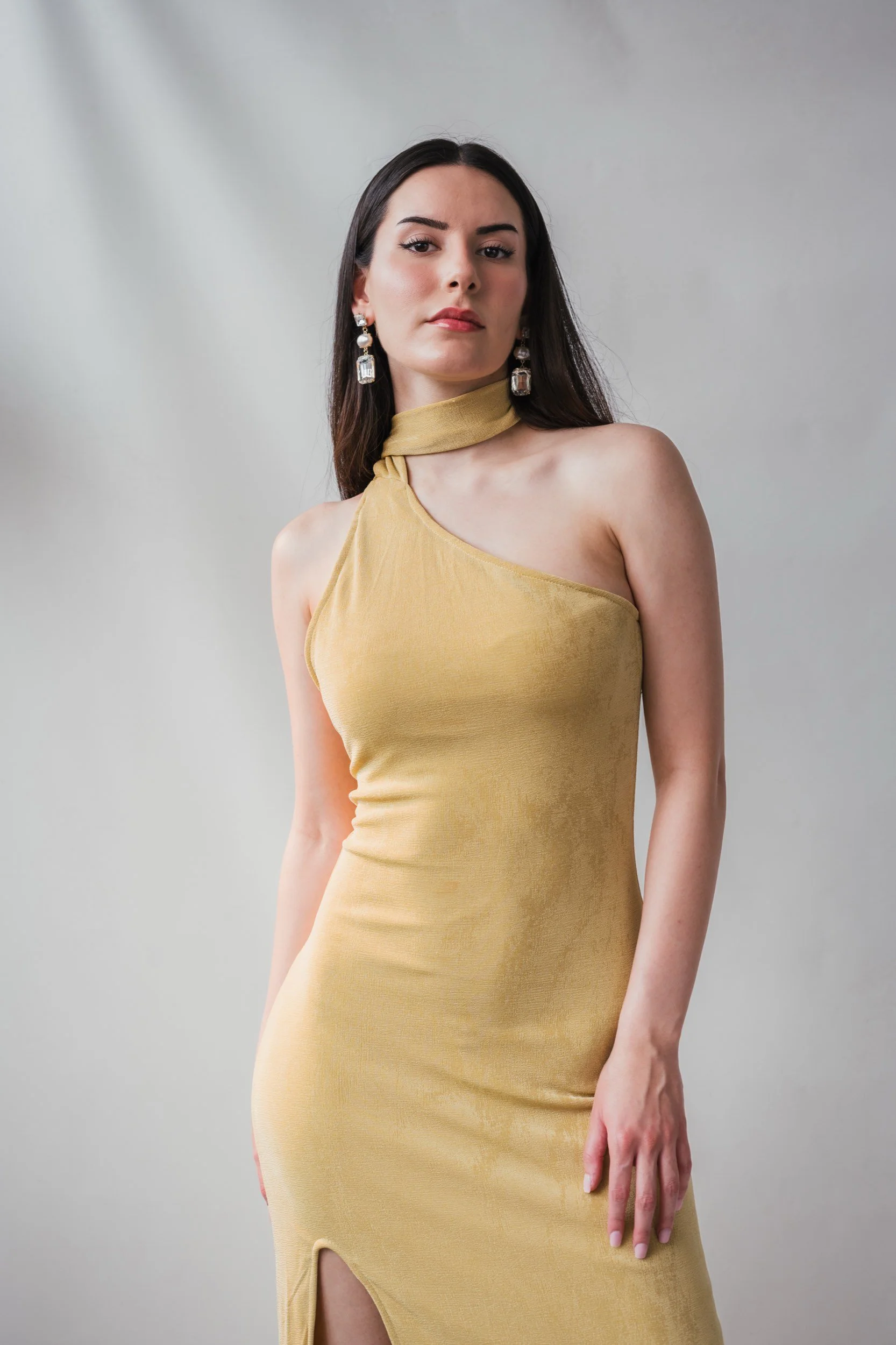 Eine junge Frau trägt ein einteiliges, schimmerndes, gelbes Kleid mit einem asymmetrischen Schulterdesign und einem hohen Schlitz. Sie hat lange dunkle Haare, trägt große, elegante Ohrringe und blickt ernst in die Kamera vor einem neutralen Hintergru
