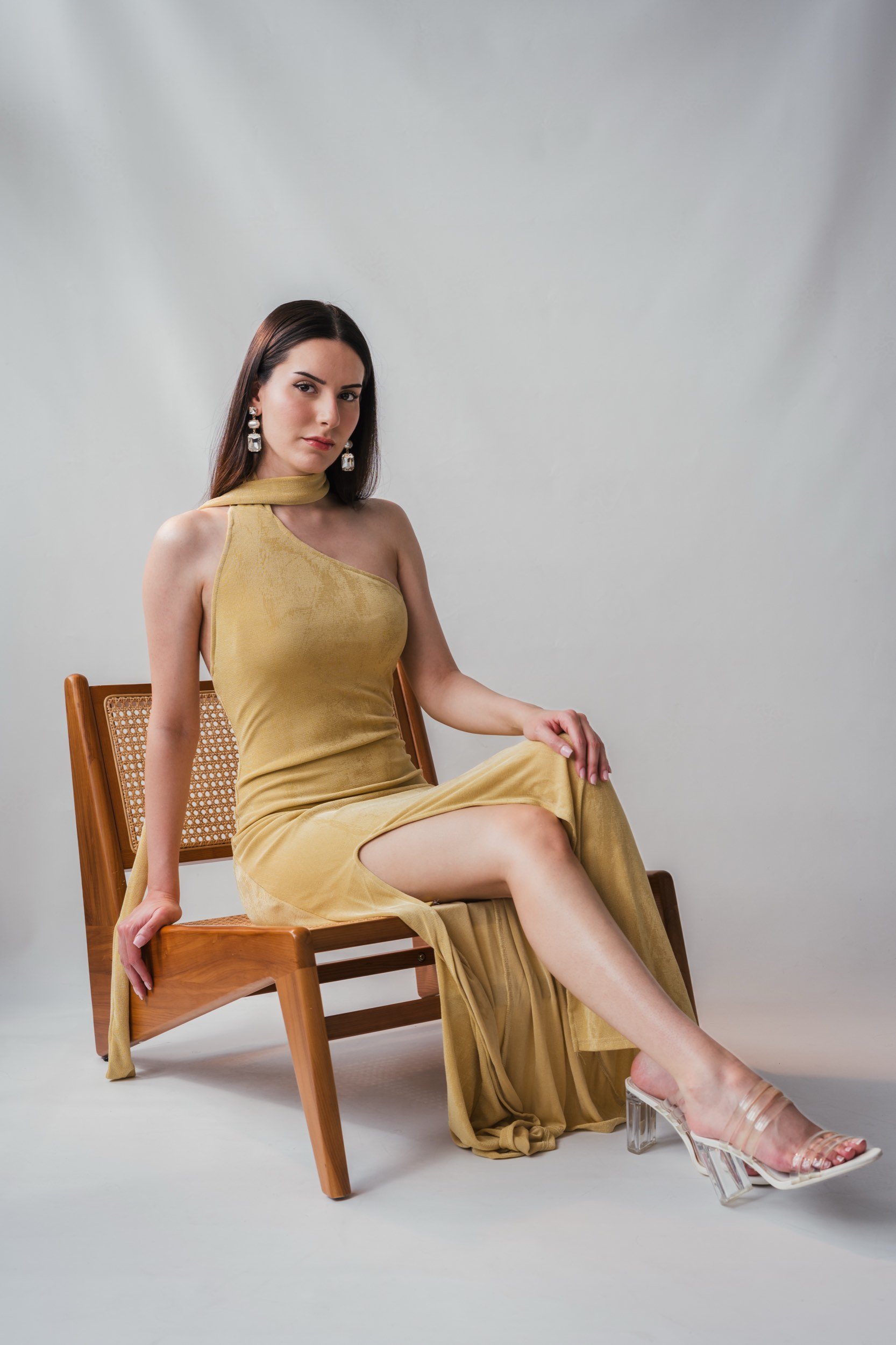 Eine Frau in einem goldgelben Kleid sitzt auf einem Holzstuhl vor weißem Hintergrund.