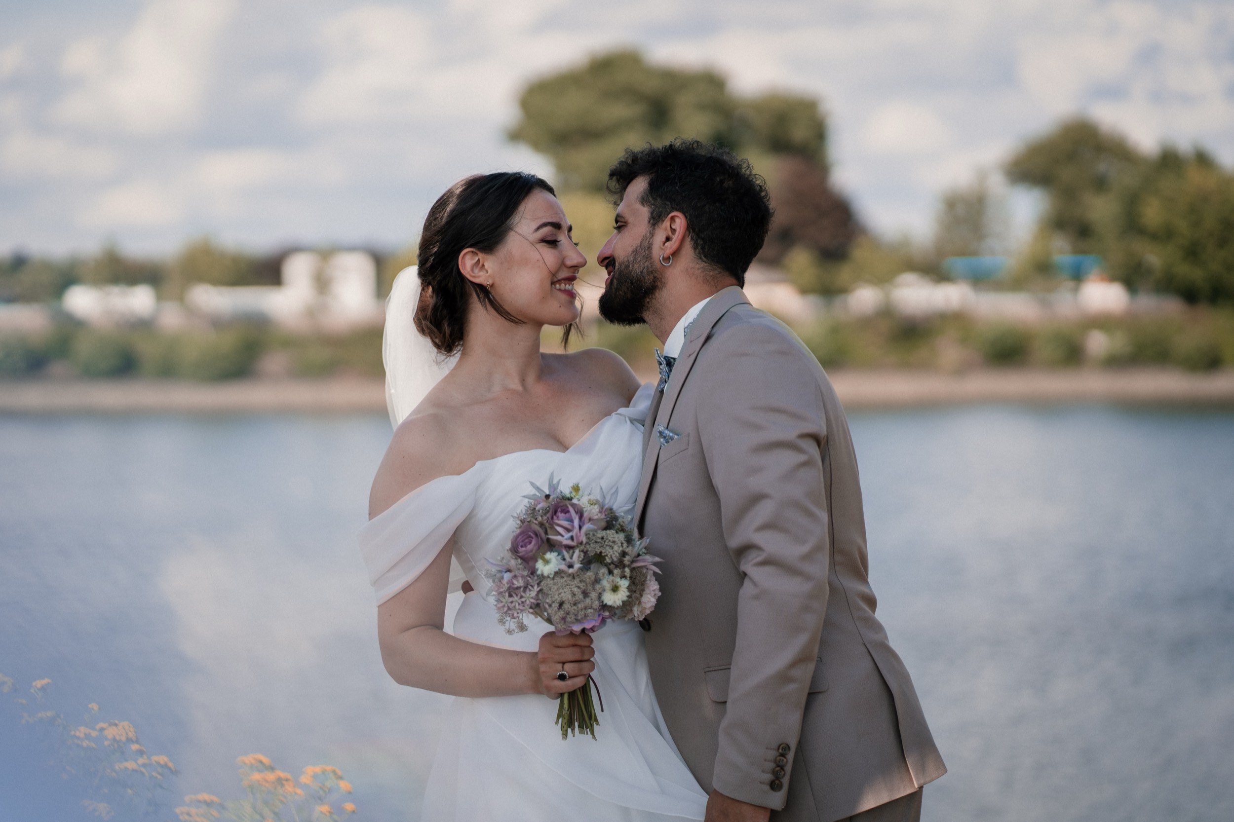 Ein verliebtes Paar bei einer Hochzeitsaufnahme am Wasser, die Frau in einem weißen Hochzeitskleid mit Brautstrauß, der Mann in einem hellgrauen Anzug. Im Hintergrund Wasser und Bäume.
