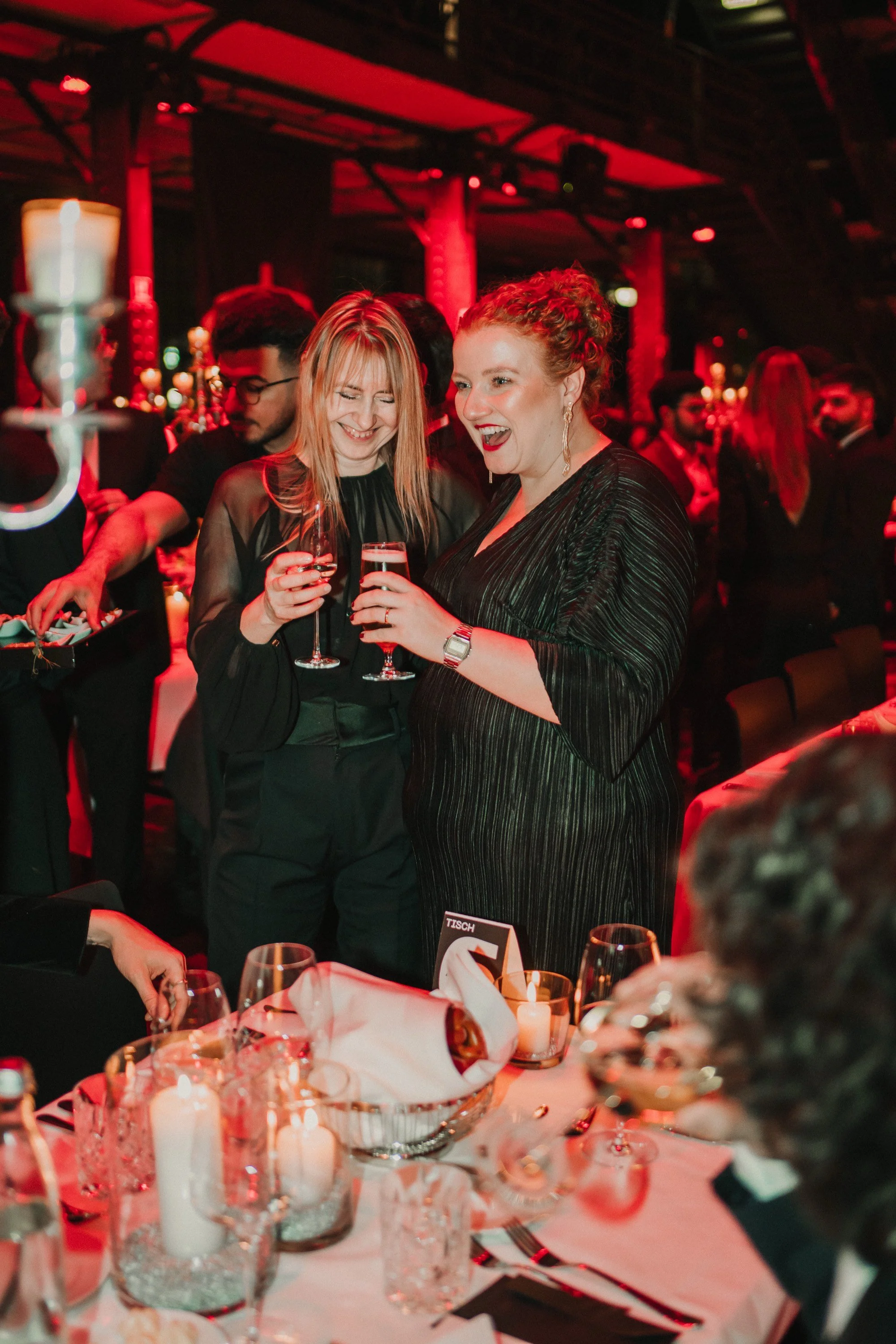 Zwei lachende Frauen, die ein Glas Wein in einer festlichen Umgebung trinken, umgeben von mehr Menschen und Kerzen auf einem Tisch bei einer Gala oder einer Party.