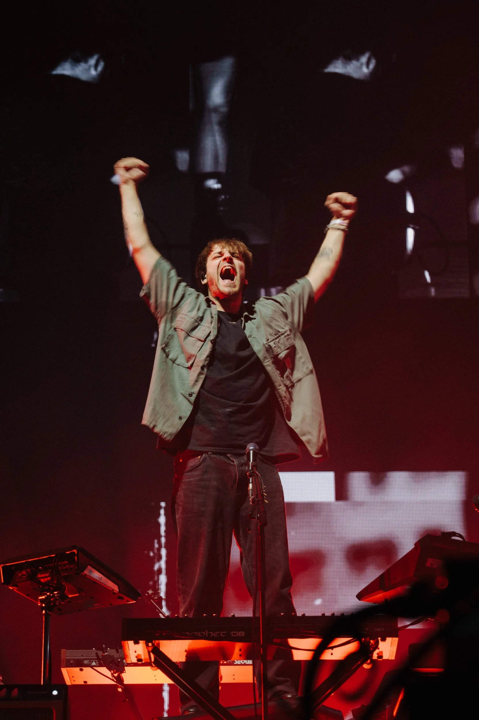 Ein männlicher Musiker steht auf einer Bühne, hebt die Arme in die Luft und schreit. Er trägt eine grüne Jacke und schwarze Kleidung, umgeben von rotem Licht und Bühnenoutlines.