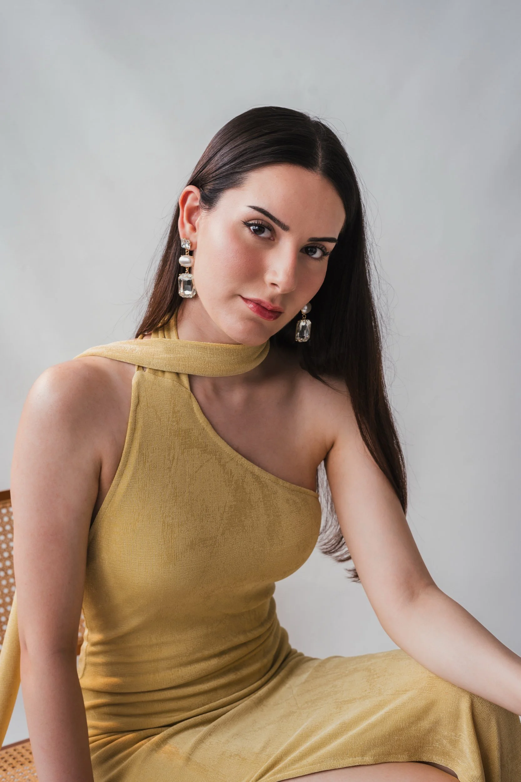 Eine junge Frau in einem goldenen ein- schulterigen Kleid mit großen Ohrringen sitzt auf einem Stuhl.