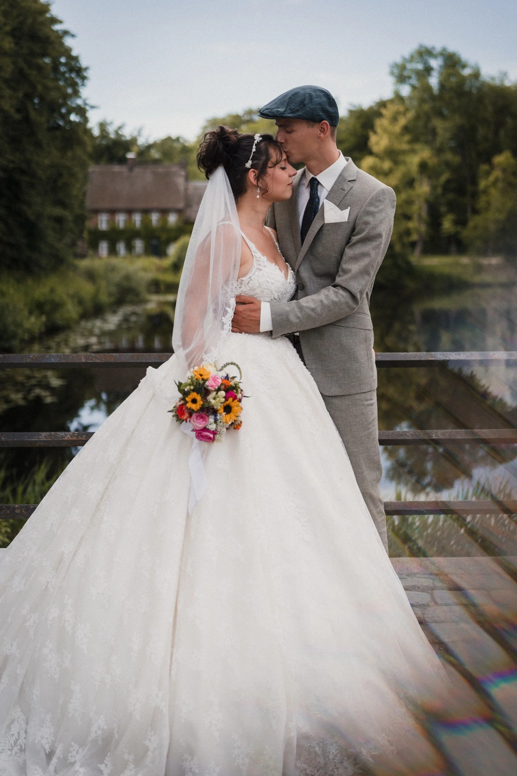 Ein Brautpaar auf einer Brücke, die Braut trägt ein weißes Brautkleid und hält einen bunten Blumenstrauß, der Bräutigam trägt einen grauen Anzug und ein Karo-Hut, im Hintergrund sind Bäume und ein Fluss zu sehen.