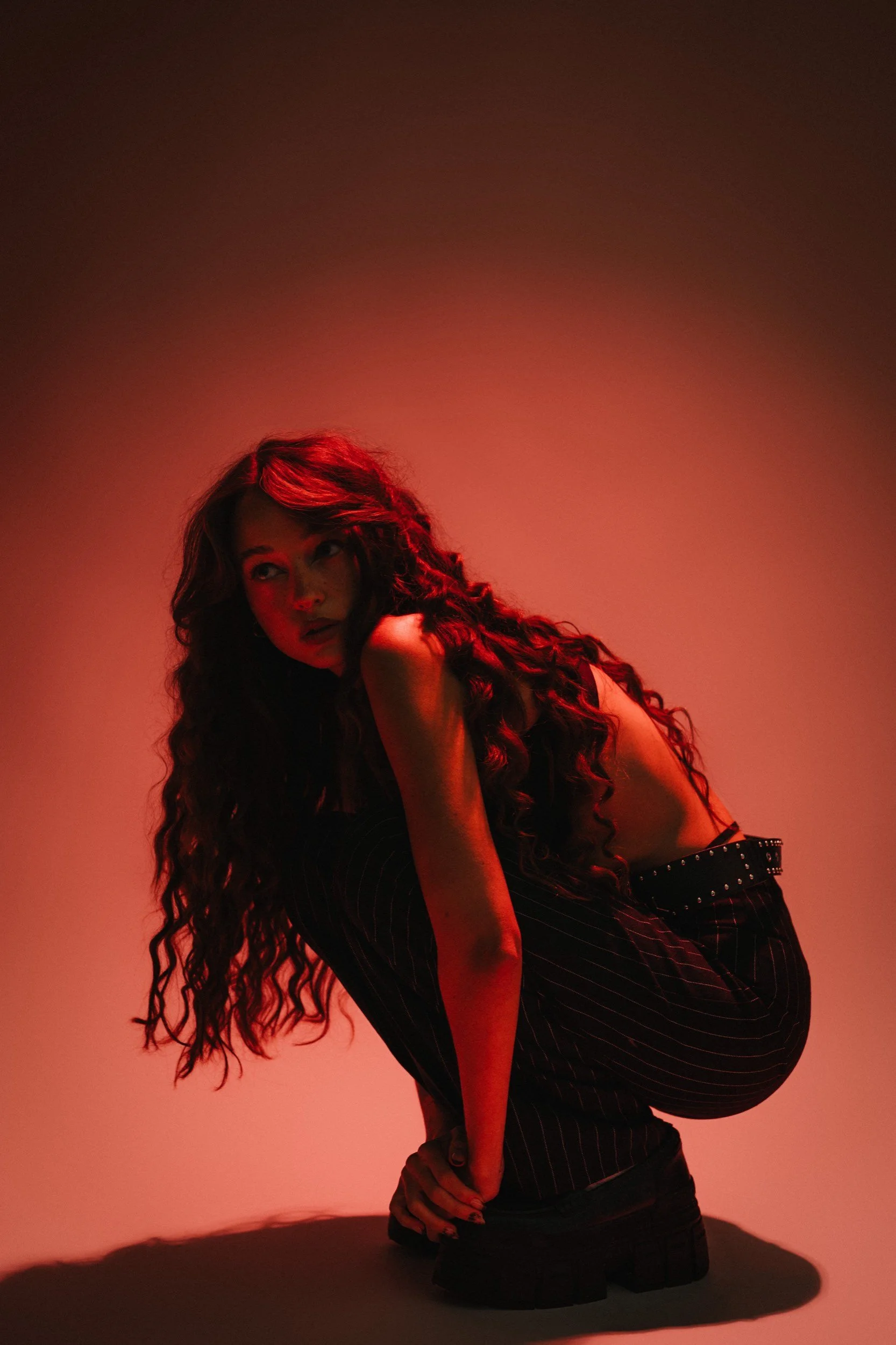 Junge Frau mit langen, lockigen Haaren in dunkler Kleidung, in einer roten Beleuchtungskulisse gekniet.