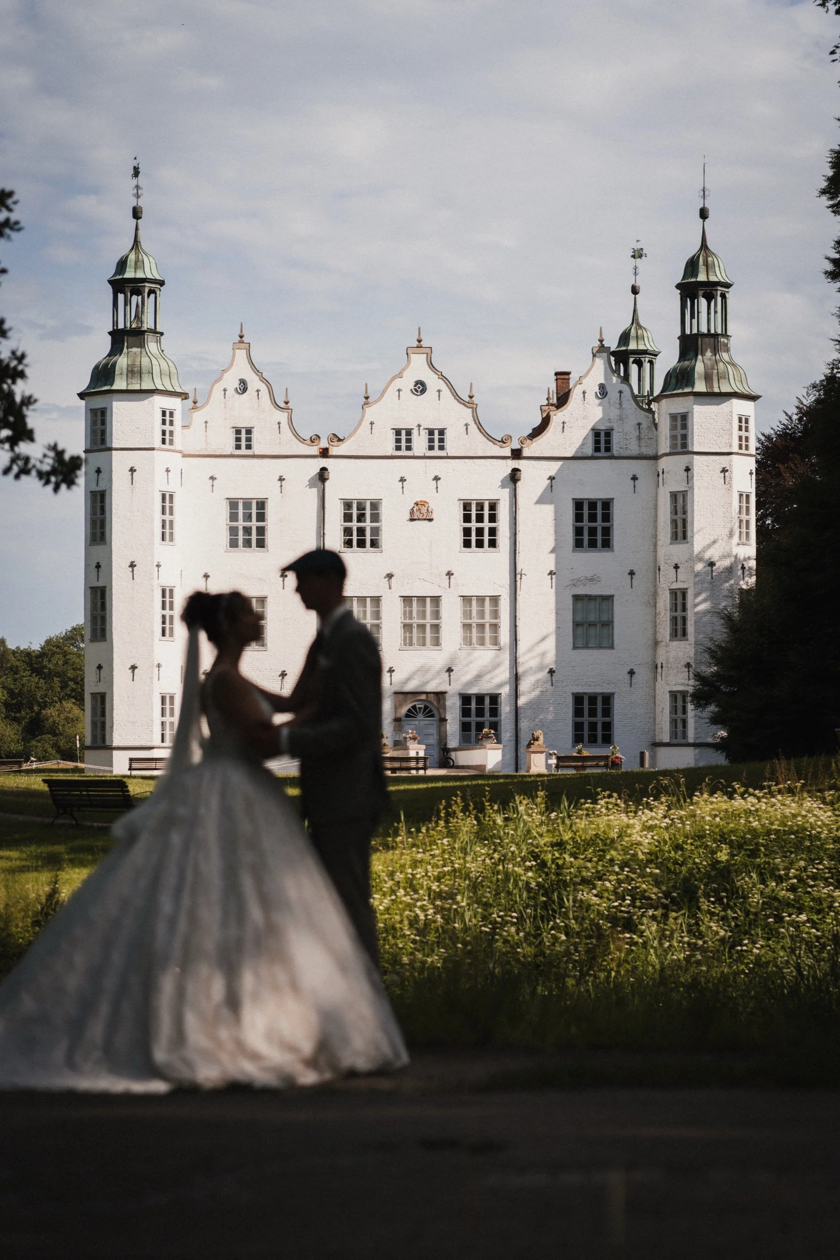 Silhouette eines Brautpaares beim Tanzen vor einem weißen Schloss mit Türmen bei Sonnenuntergang.