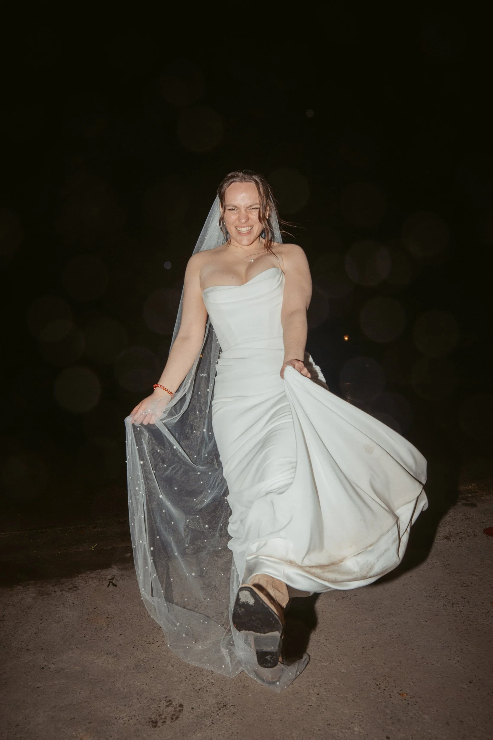 Frau in Brautkleid beim Tanzen auf einer dunklen Außenfläche mit nassem Boden bei Nacht.