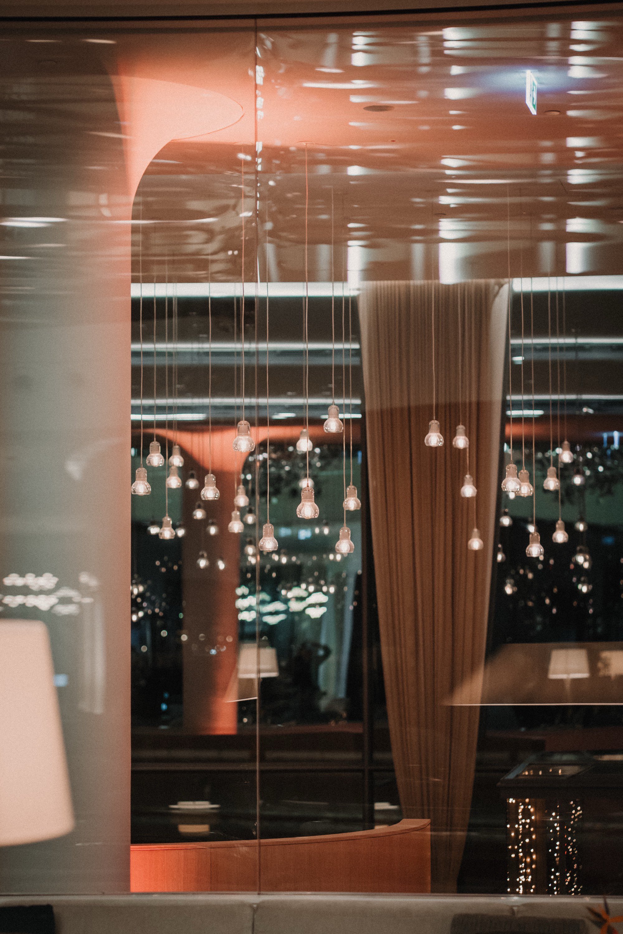 Innenaufnahme eines modernen, beleuchteten Raums mit Hängelampen und großen Fenstern, durch die die Stadt bei Nacht sichtbar ist.