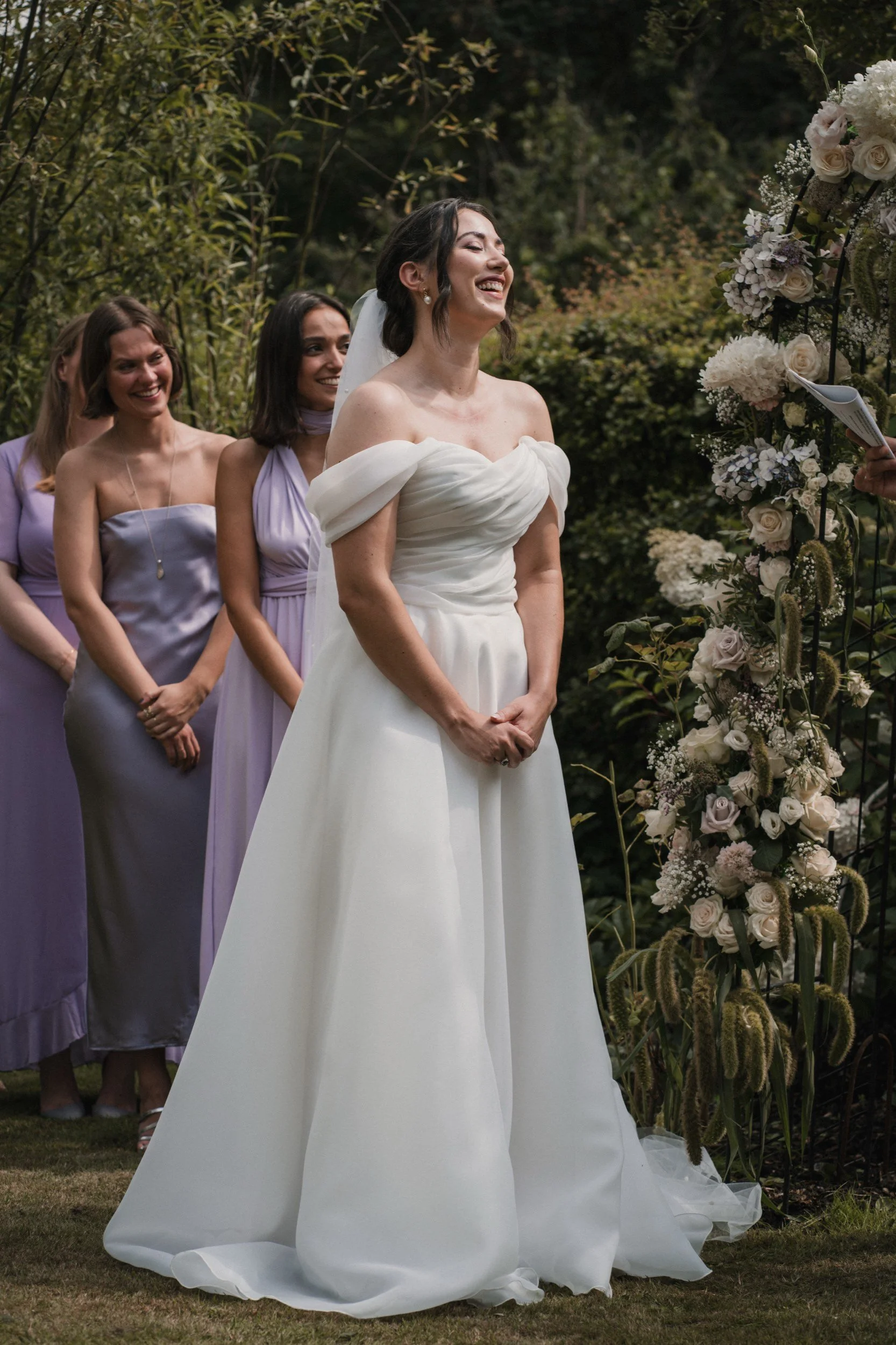Eine glückliche Frau in einem weißen Brautkleid bei einer Hochzeit im Freien, umgeben von drei Frauen in lilafarbenen Kleidern, mit einer Blumenwand im Hintergrund.
