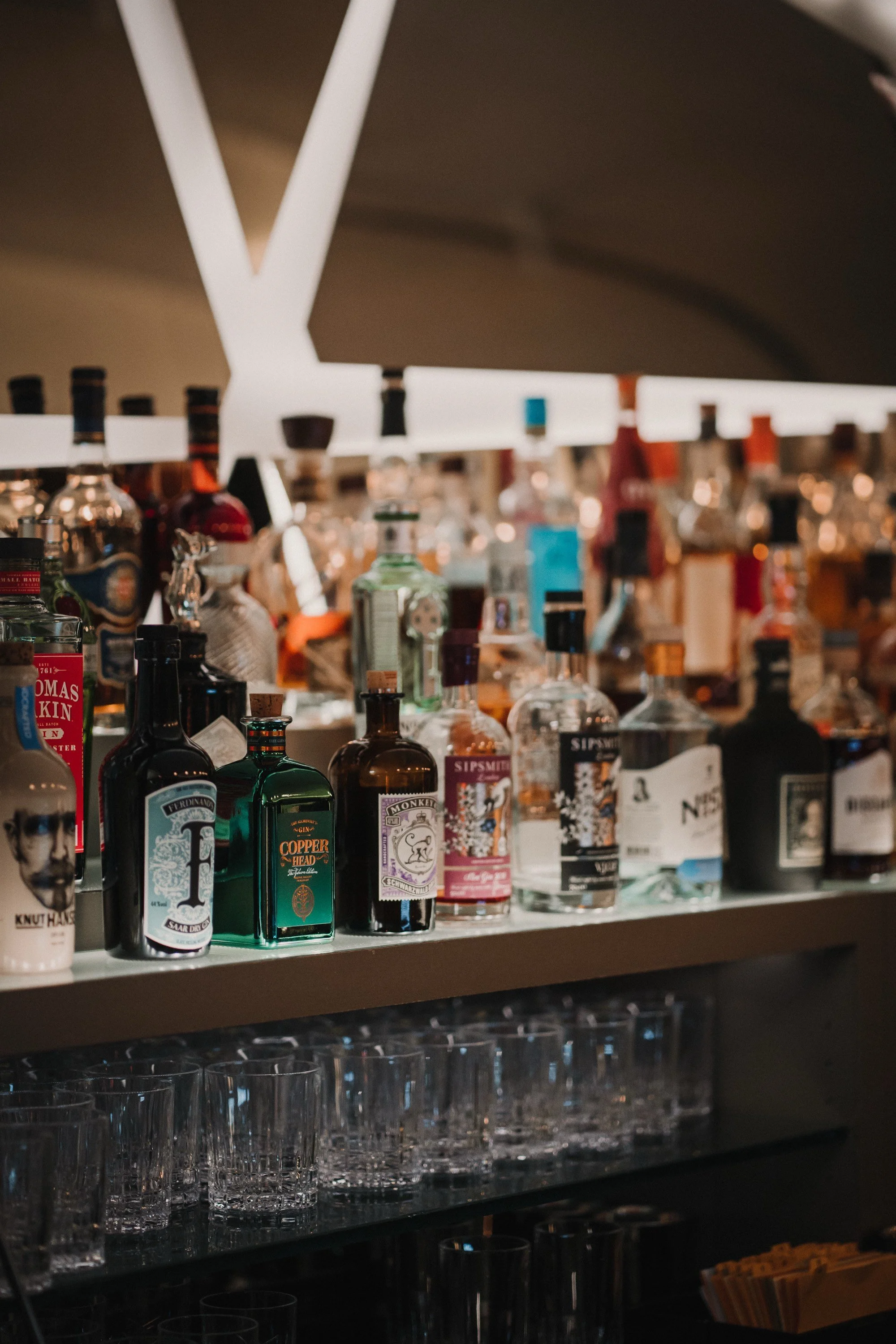 Verschiedene Spirituosenflaschen auf einer Bar-Regal, darunter Gin, Wodka und Liköre, mit leeren Gläsern darunter.