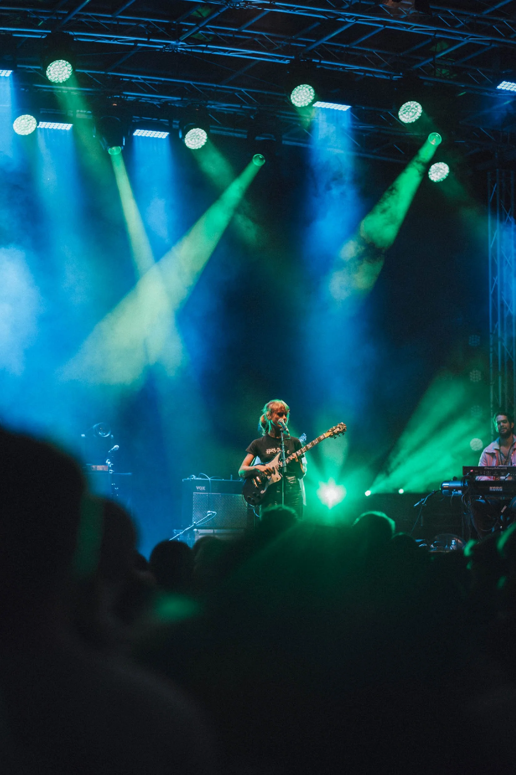 Eine Musikerin spielt Gitarre auf einer Bühne bei einem Konzert, umgeben von bunten Scheinwerfern und einer dunklen Kulisse.