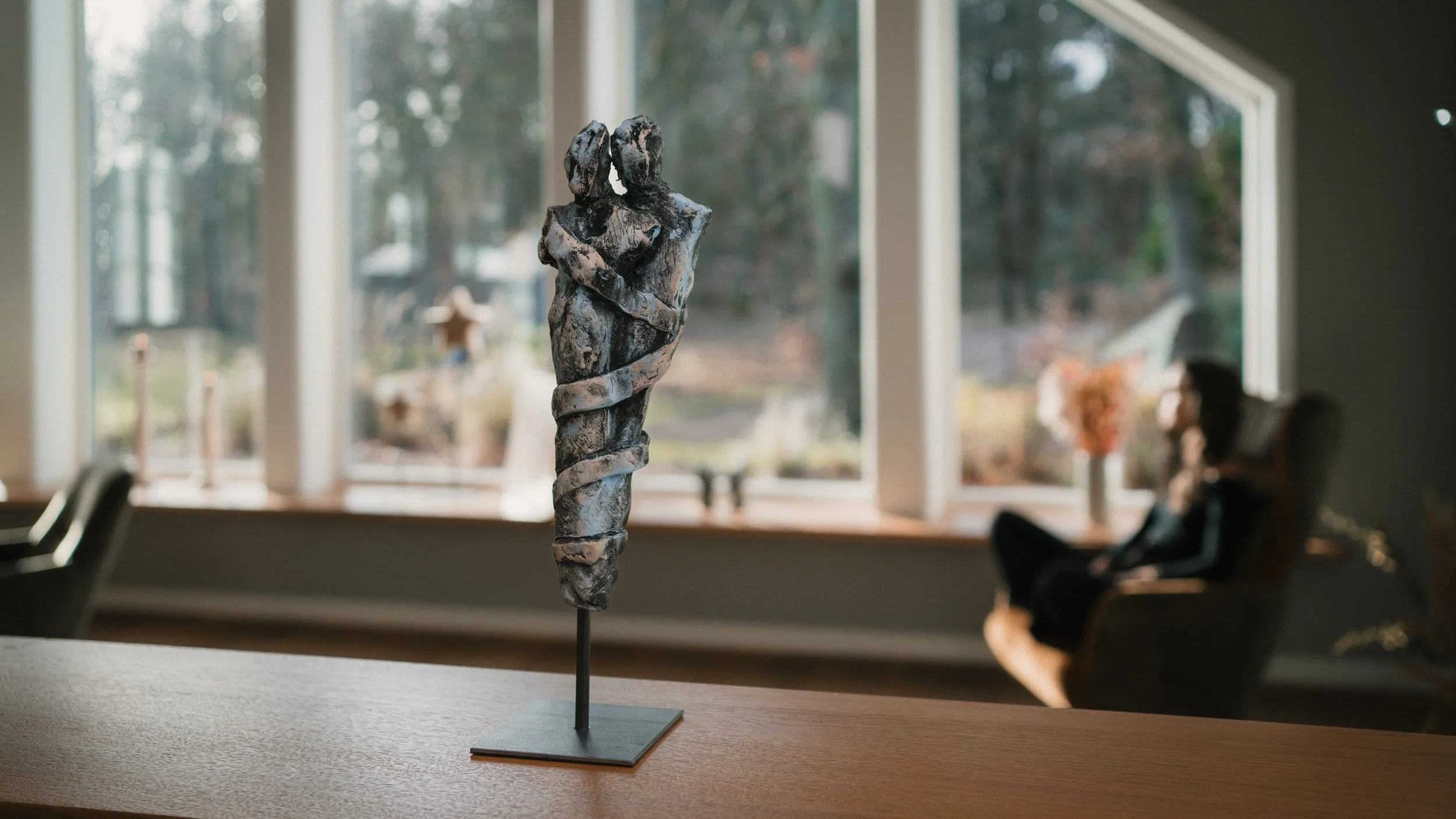 Kunstwerk einer Skulptur eines Paares, das sich umarmt, auf einem Tisch vor einem Fenster, mit einer Frau, die im Hintergrund sitzend den Blick nach draußen richtet.