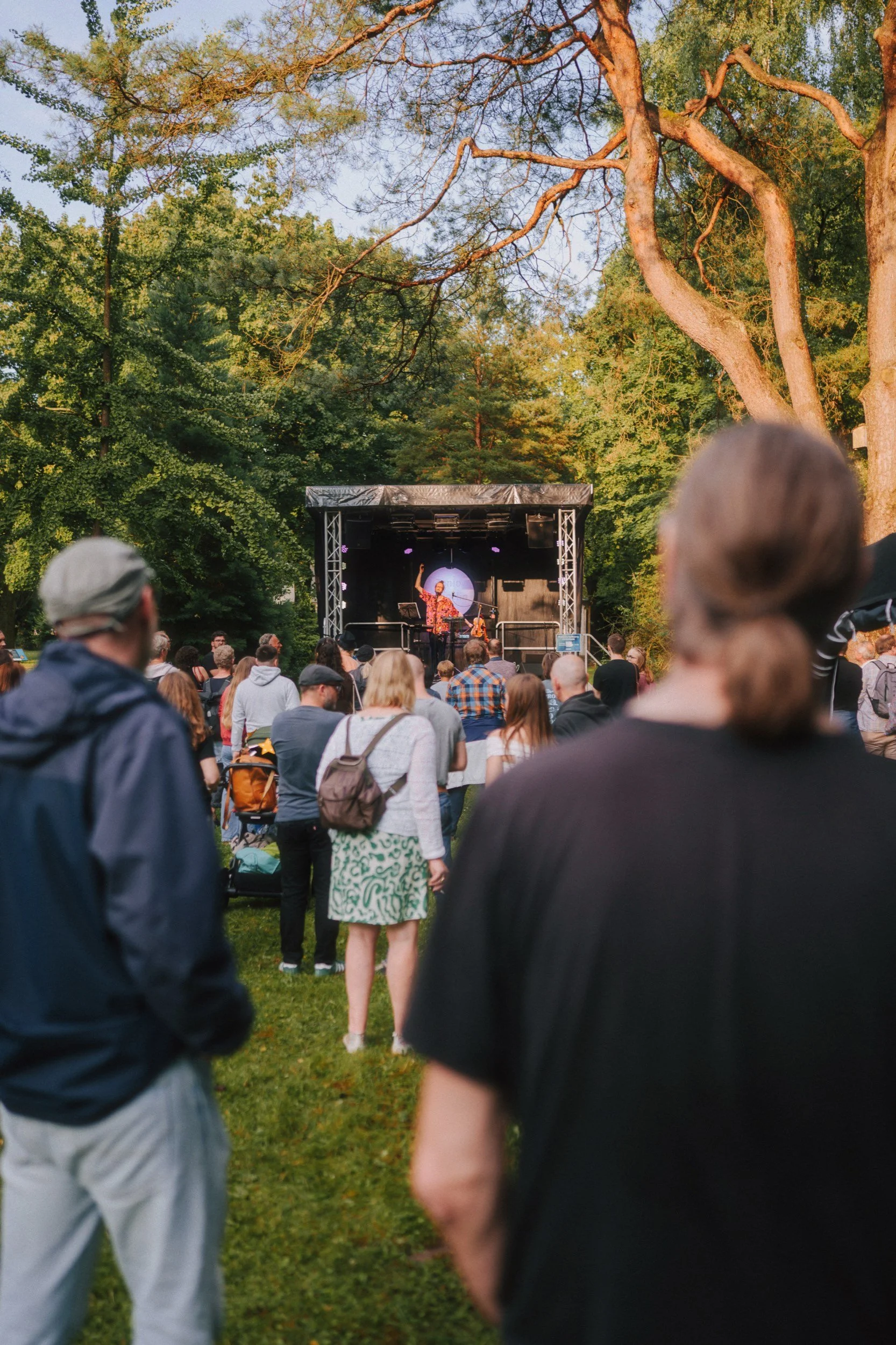 Menschen, die einem Konzert im Freien auf einer Bühnen im Park zuhören, umgeben von Bäumen bei Sonnenuntergang.