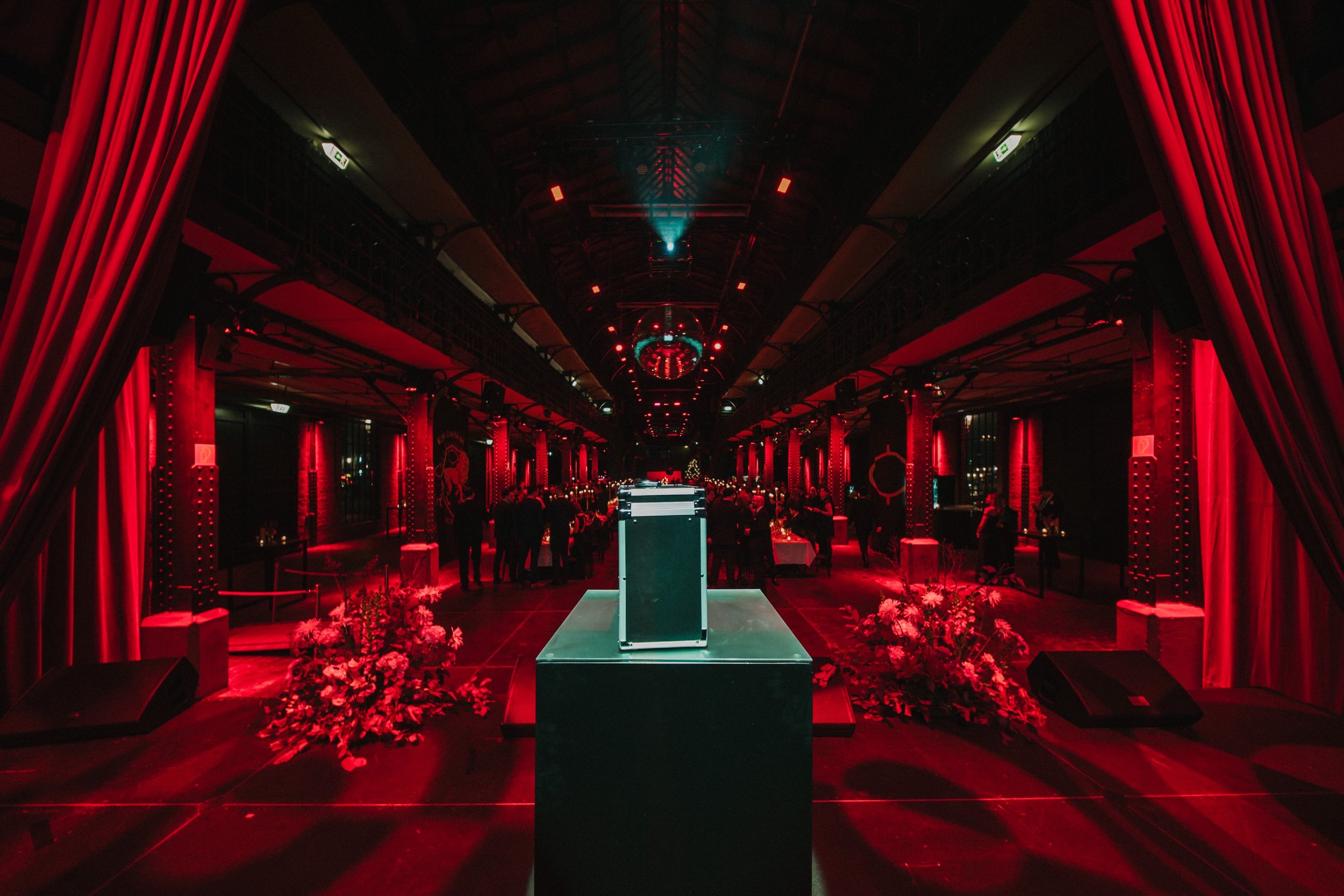 Bühne für eine Veranstaltung mit rotem Beleuchtung, Blumenarrangements und Gästen im Hintergrund.
