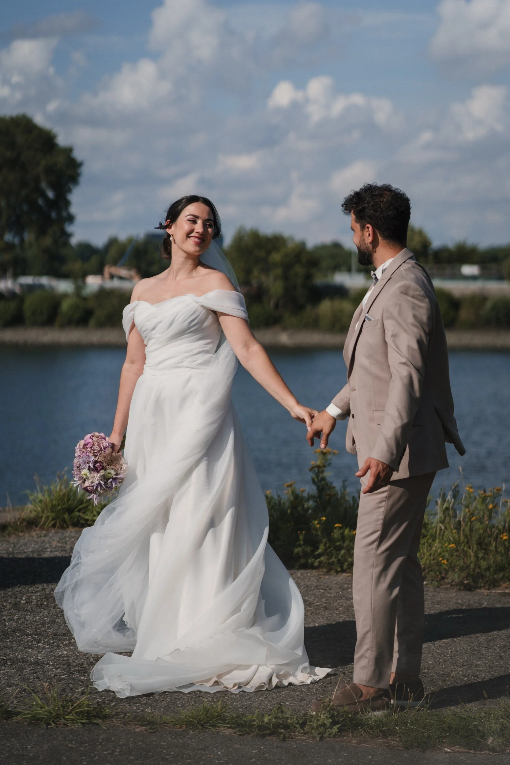 Ein verlobtes Paar hält Händchen an einem Wasserufer, die Frau trägt ein weißes Brautkleid und hält einen Blumenstrauß, der Mann trägt einen hellbeigen Anzug.