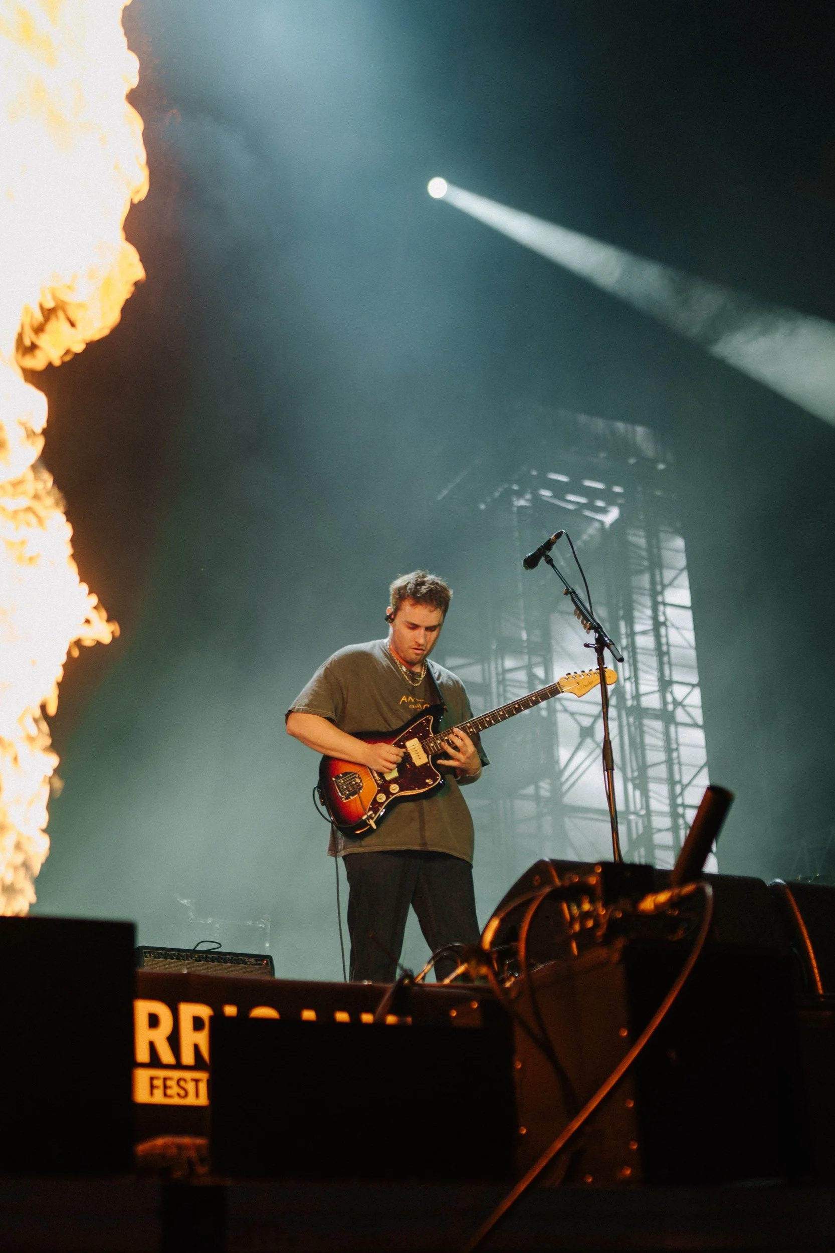 Ein junger Mann spielt Gitarre auf einer Bühne bei einem Live-Konzert, im Hintergrund wirft Feuer Flammen in die Luft, während ein starker Lichtstrahl auf die Bühne scheint.