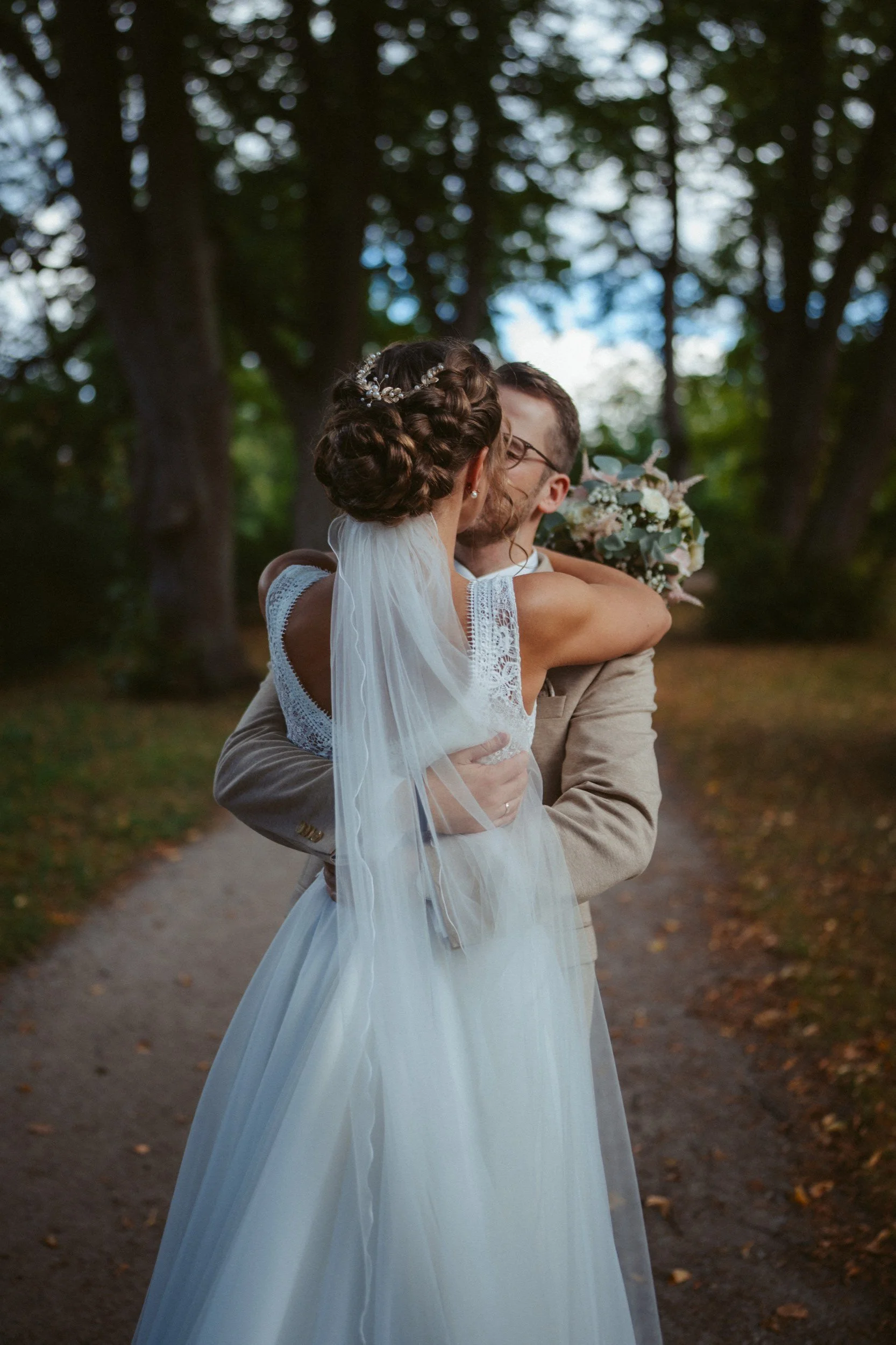 Ein Brautpaar umarmt sich auf einem Waldweg bei Sonnenuntergang, die Braut trägt ein weißes Hochzeitskleid mit langer Seidenschleier, der Bräutigam trägt einen hellen Anzug.