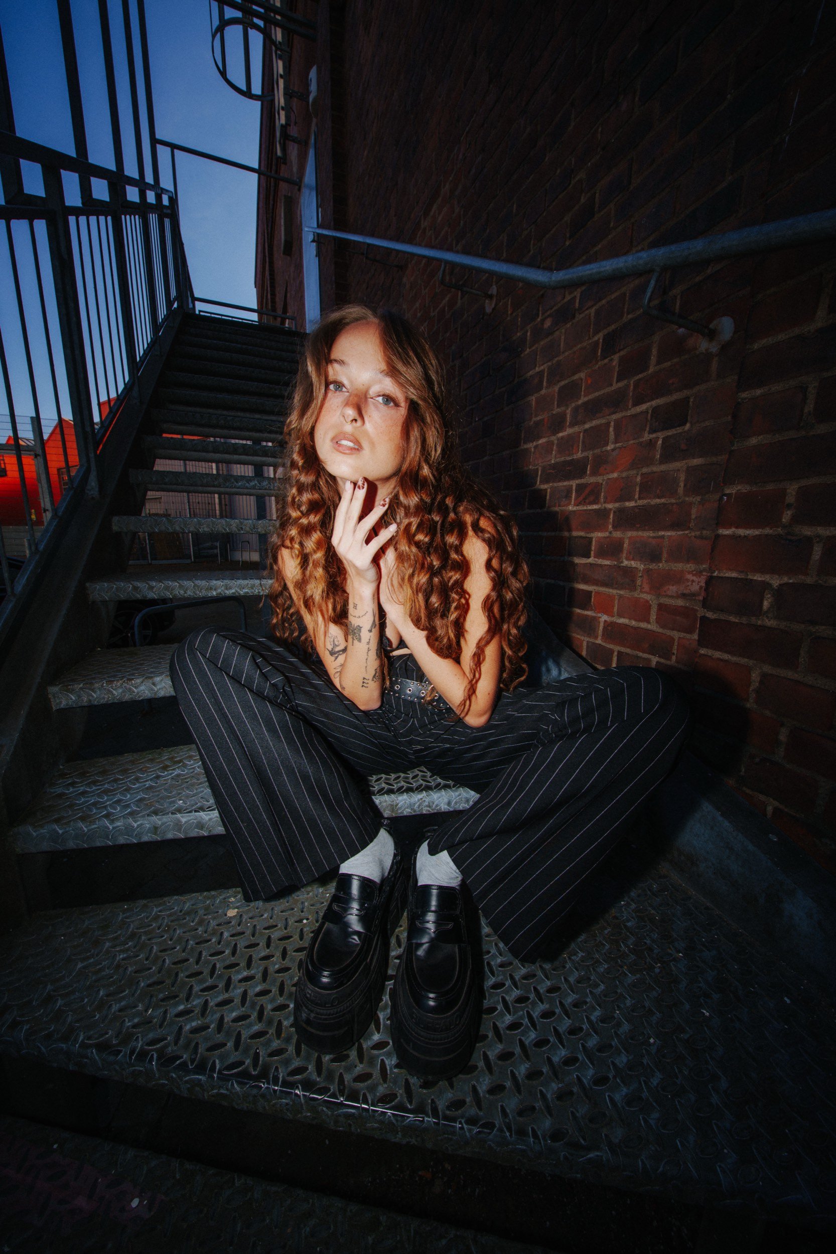 Junge Frau mit langen, lockigen braunen Haaren sitzend auf einer Metalltreppe auf einem Balkon im Abendlicht in urbanem Umfeld