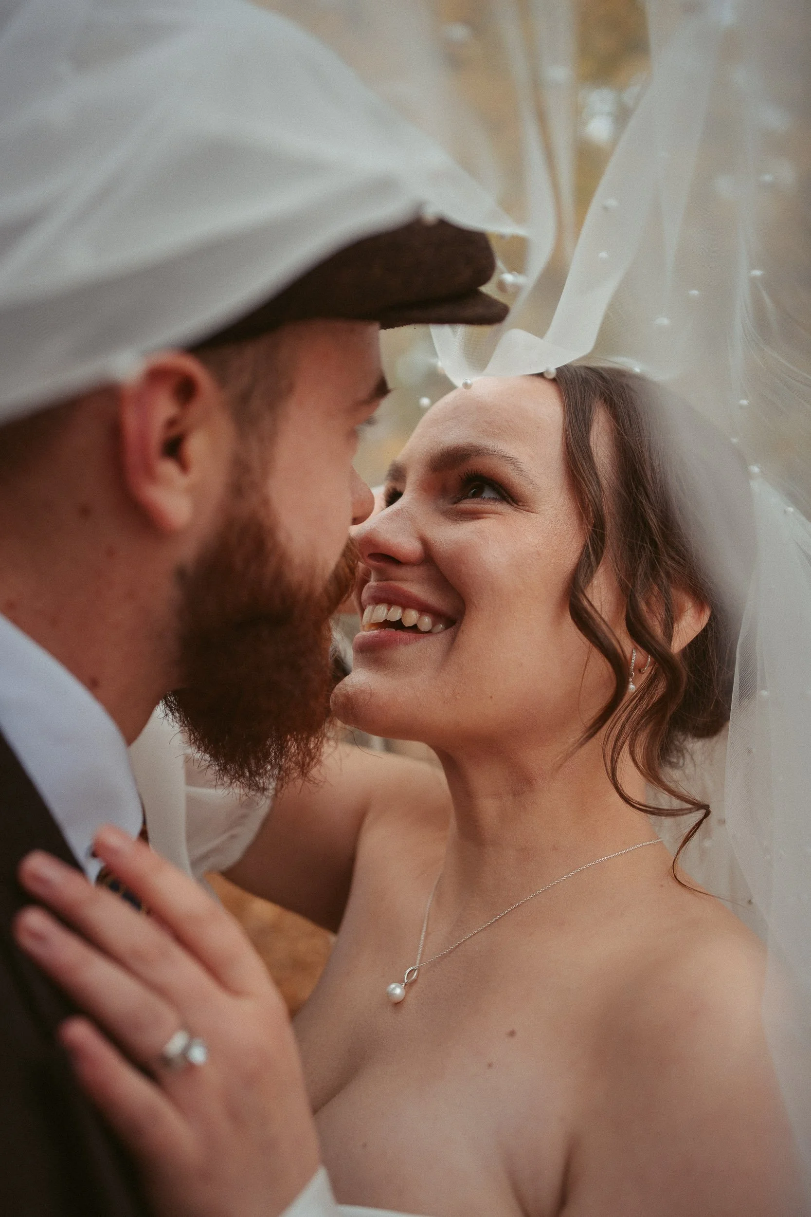 Ein braunhaariges, lachendes Brautpaar bei der Hochzeitsfeier, eng umarmt und lächelnd, im Freien mit Herbstlaub im Hintergrund.
