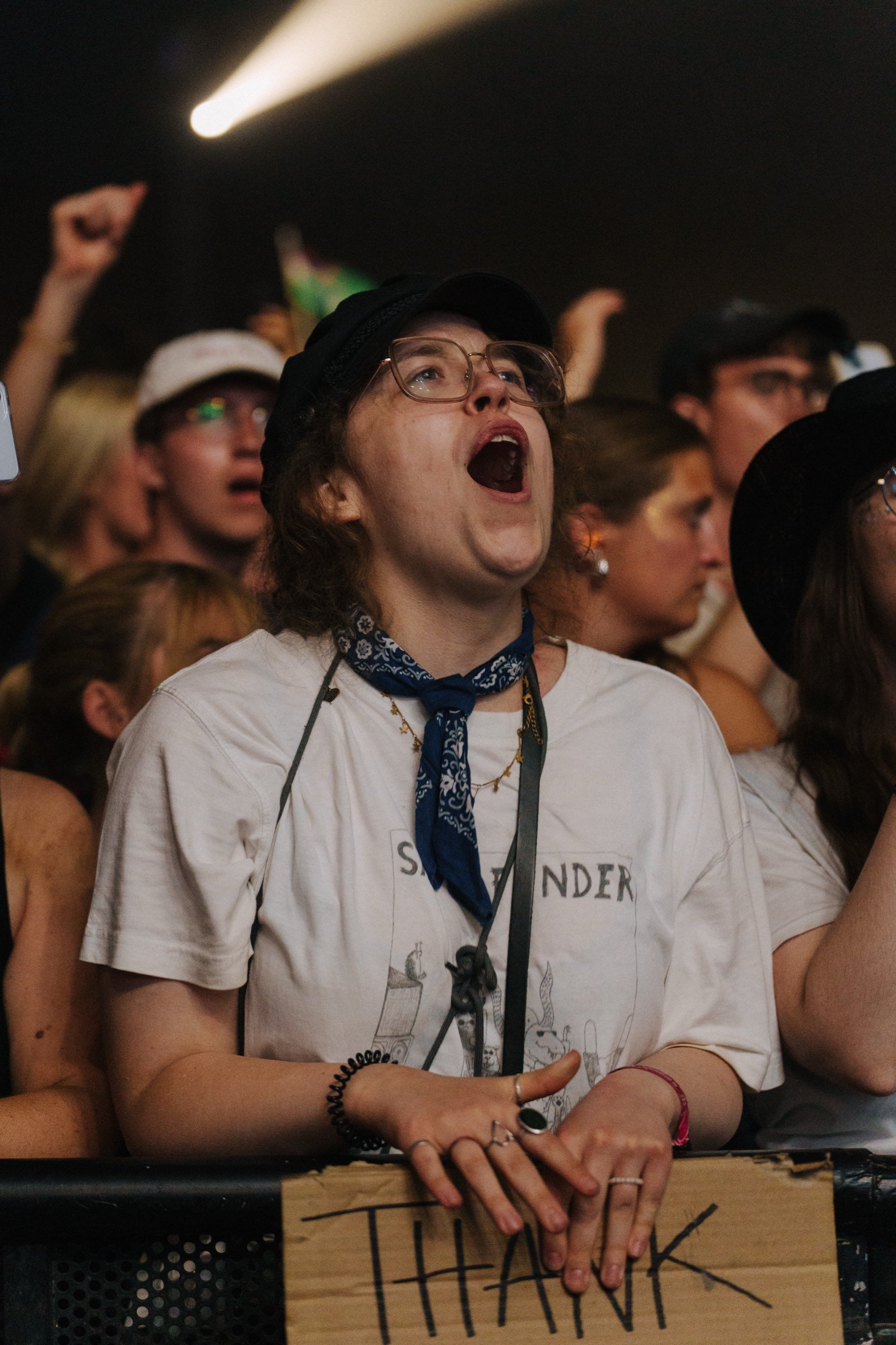 Junge Frau bei einem Konzert, die enthusiastisch singt oder ruft, mit anderen Menschen im Hintergrund, die ebenfalls anfeuern, bei Nacht oder dunklem Raum.