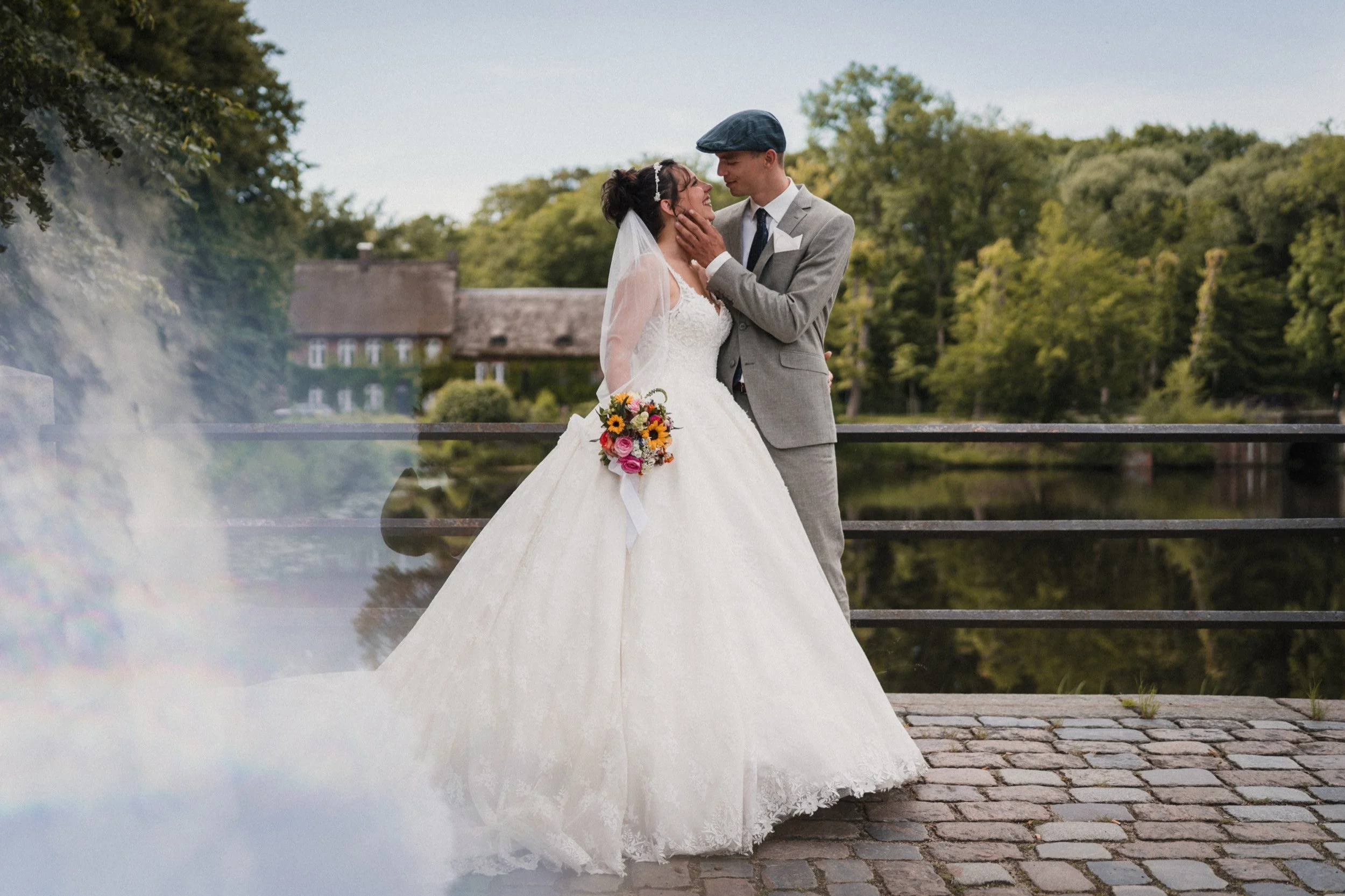 Ein Brautpaar steht am Ufer eines Flusses, die Braut trägt ein weißes Hochzeitskleid und hält einen Blumenstrauß, während der Bräutigam ein graues Anzug mit einer Baskenmütze trägt. Sie schauen sich liebevoll an.