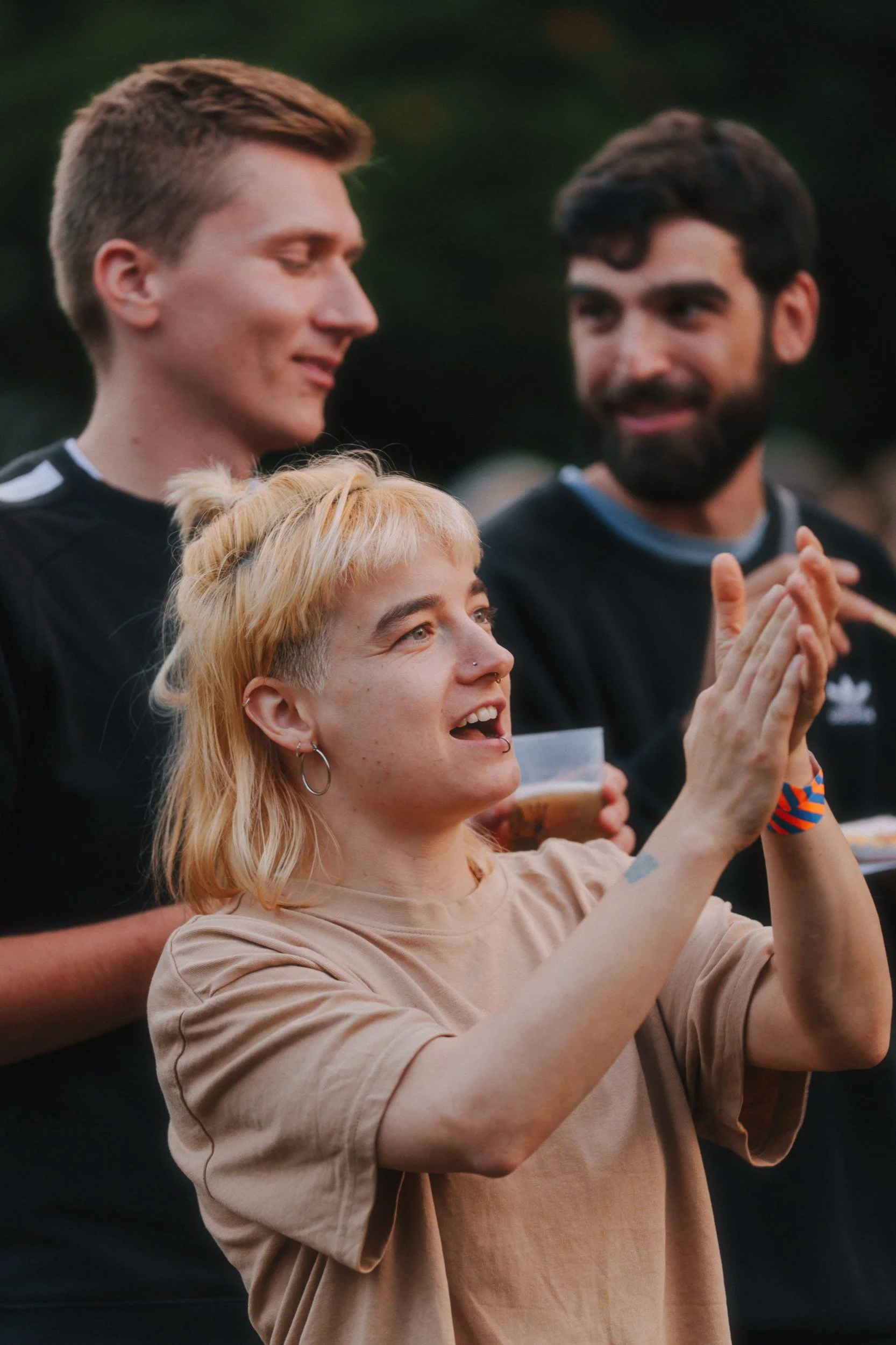 Drei junge Erwachsene feiern gemeinsam im Freien, die Frau in Vordergrund klatscht und lacht, die Männer im Hintergrund schauen und lächeln. Es ist Abenddämmerung.