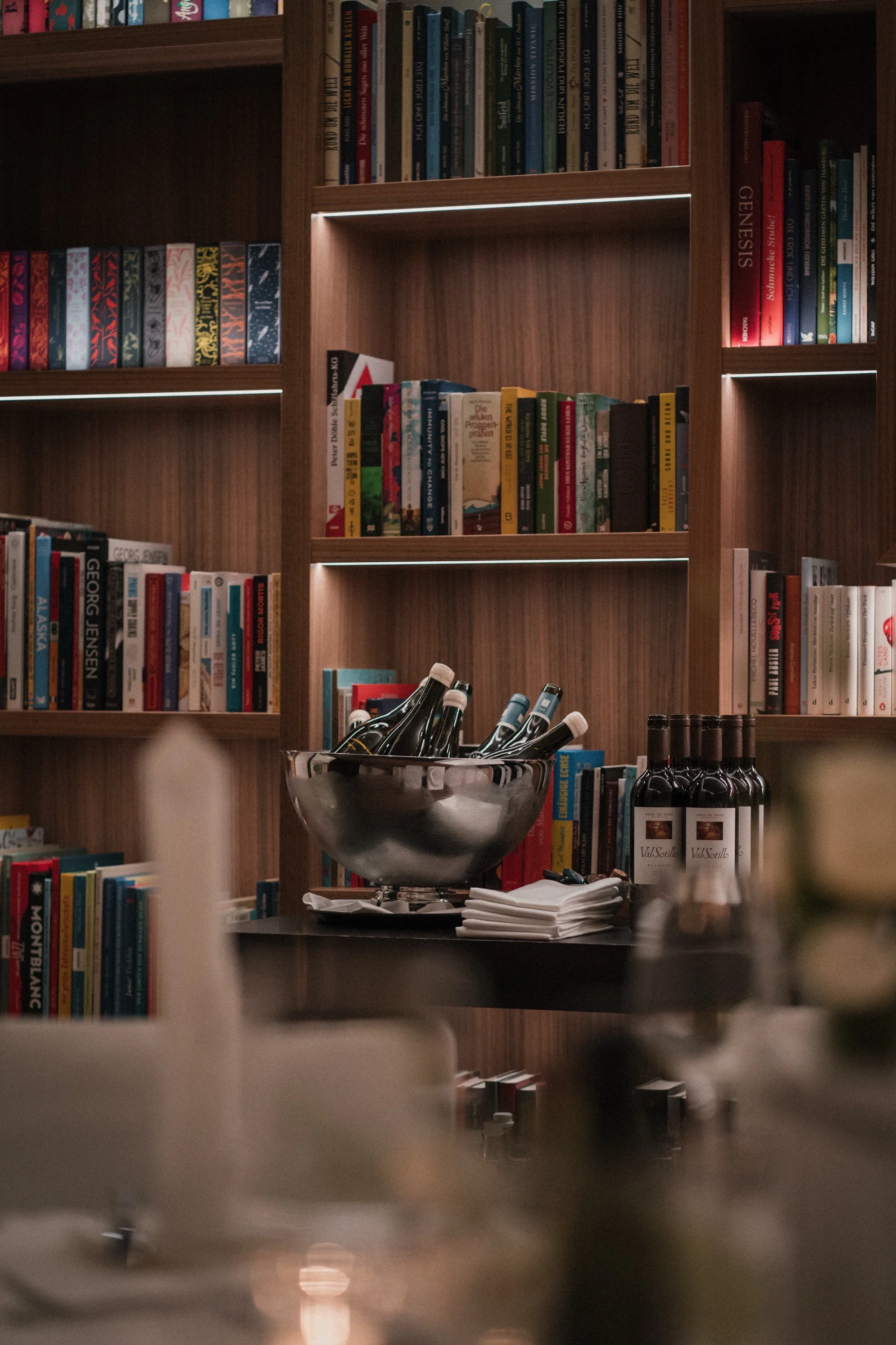 Bücherregale in einer Holzbuchhandlung oder Bibliothek, Schüsseln mit Flaschen und Flaschenöffnern, neben Weinflaschen und anderen Getränken auf einem Tisch.