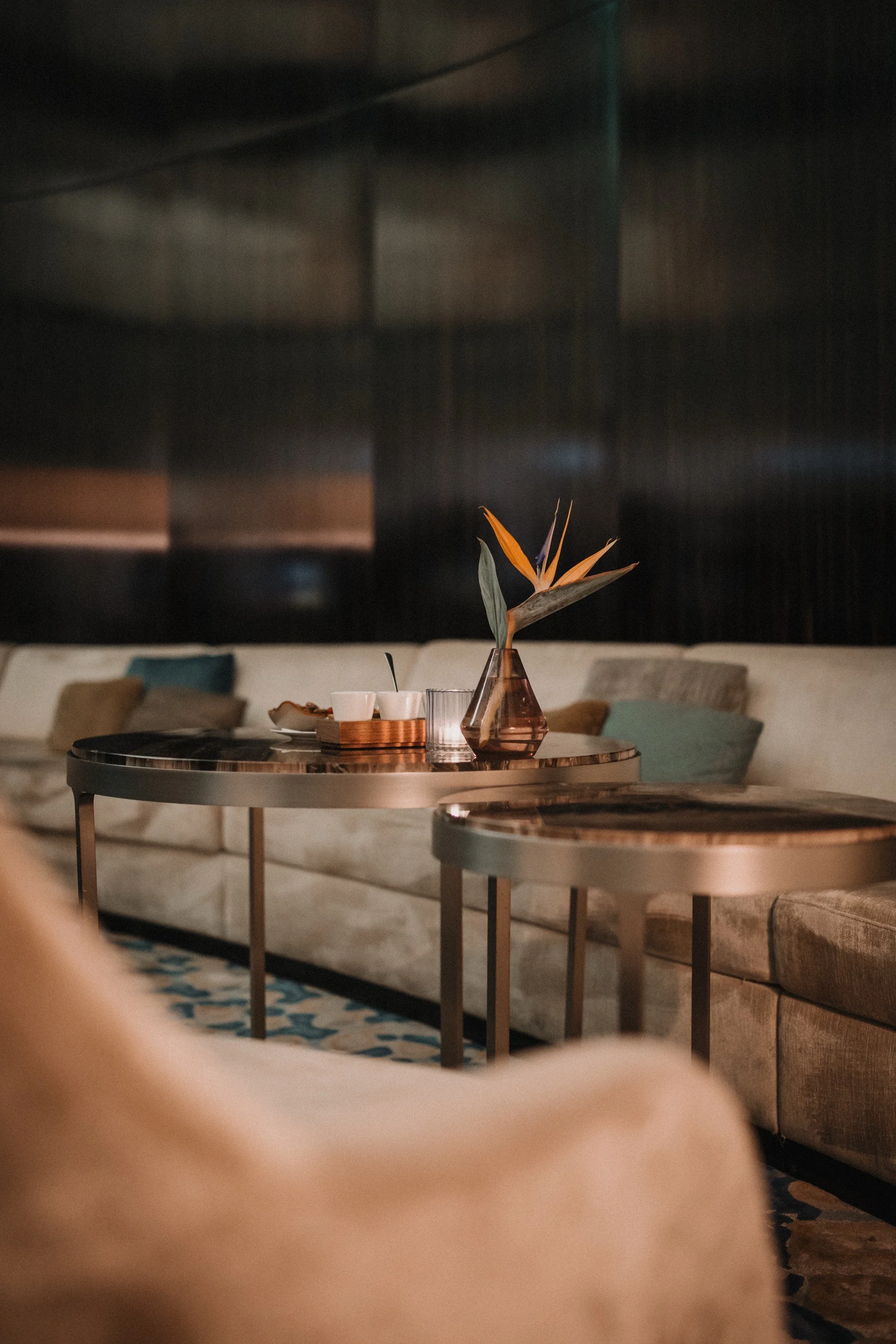 Ein gemütliches Hotel-Lounge mit einem runden Tisch, einer Vase mit tropischer Blume, Tassen und Geschirr, bequemen Sitzgelegenheiten und einem dunklen Hintergrund.