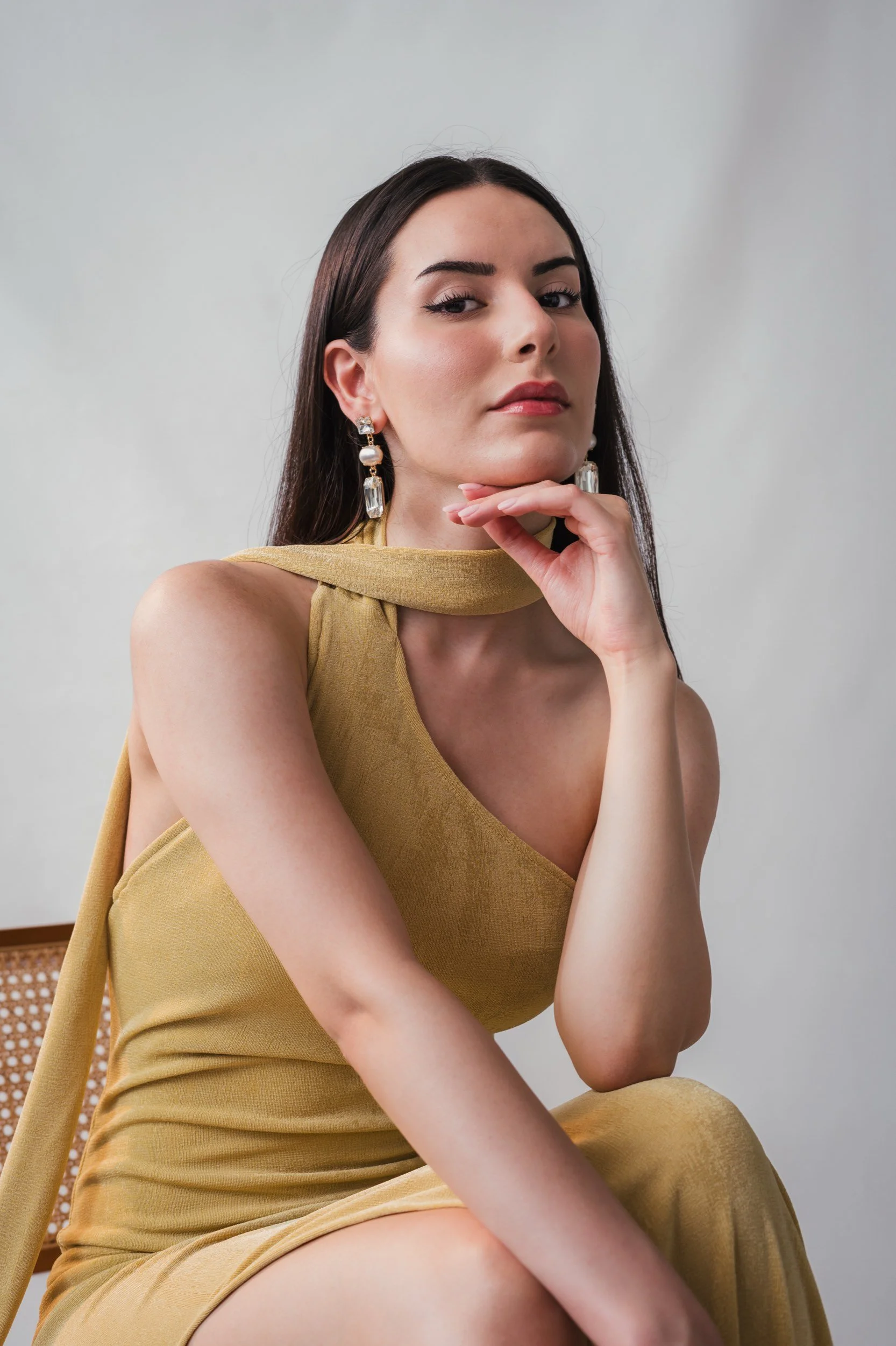 Porträt einer Frau in einem gelben Kleid mit großen Ohrringen, sitzend an einem Stuhl, schaut selbstbewusst in die Kamera.