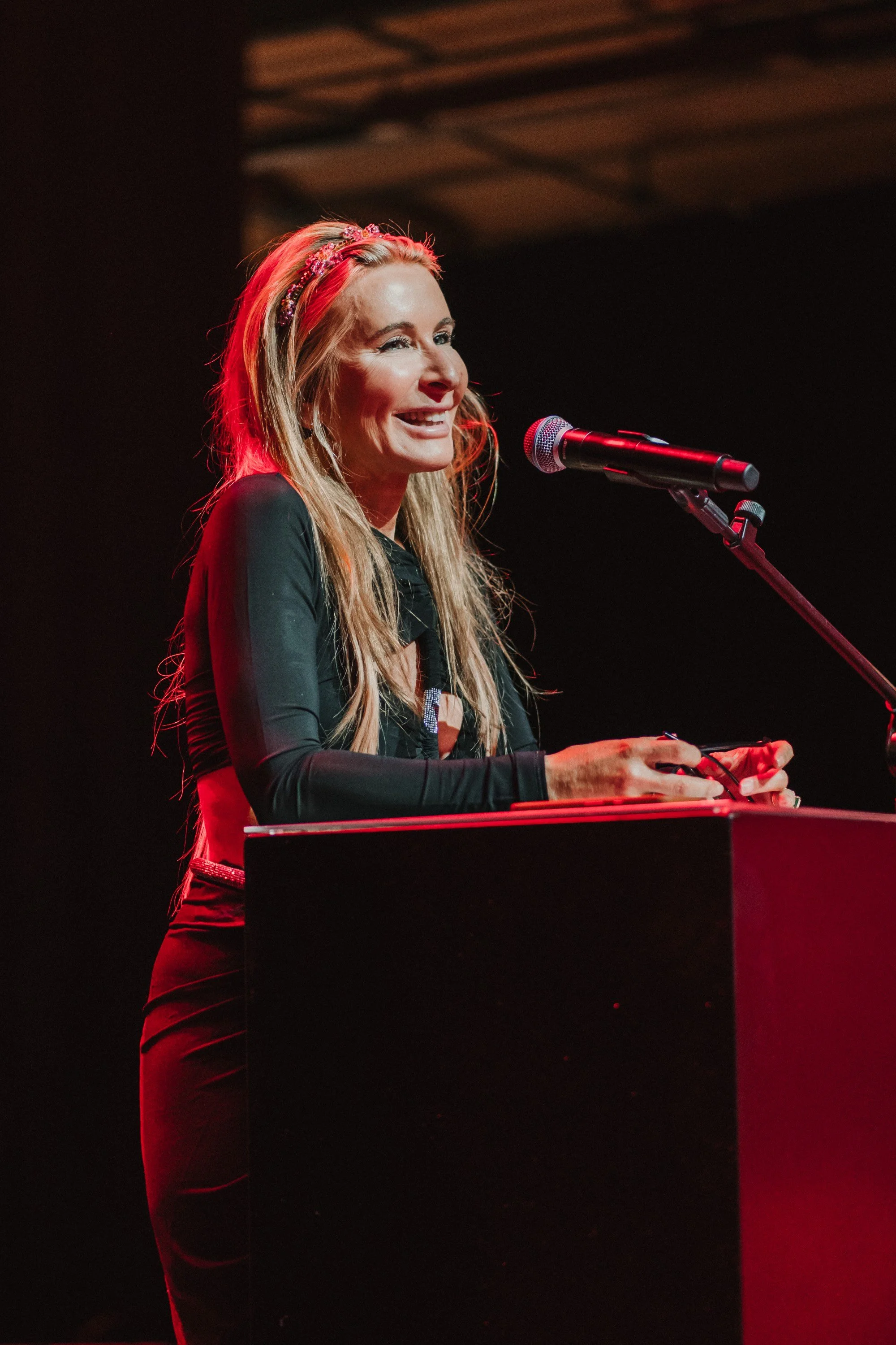 Eine lachende Frau steht an einem Pult mit einem Mikrofon, vermutlich bei einer Live-Veranstaltung oder einem Auftritt.