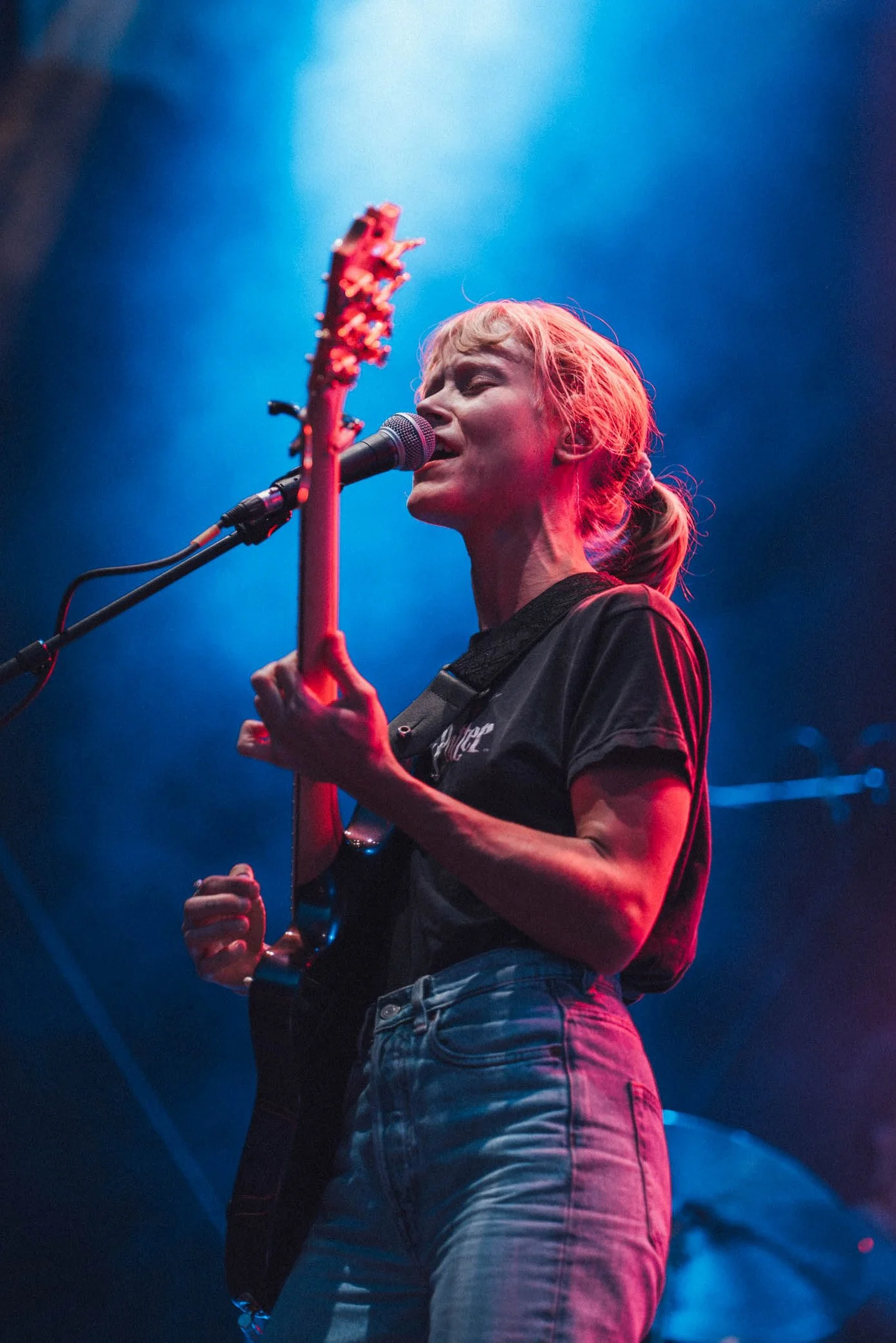 Junge Frau singt und spielt Gitarre auf einer Bühne bei einem Live-Konzert, mit blauen und roten Lichteffekten im Hintergrund.