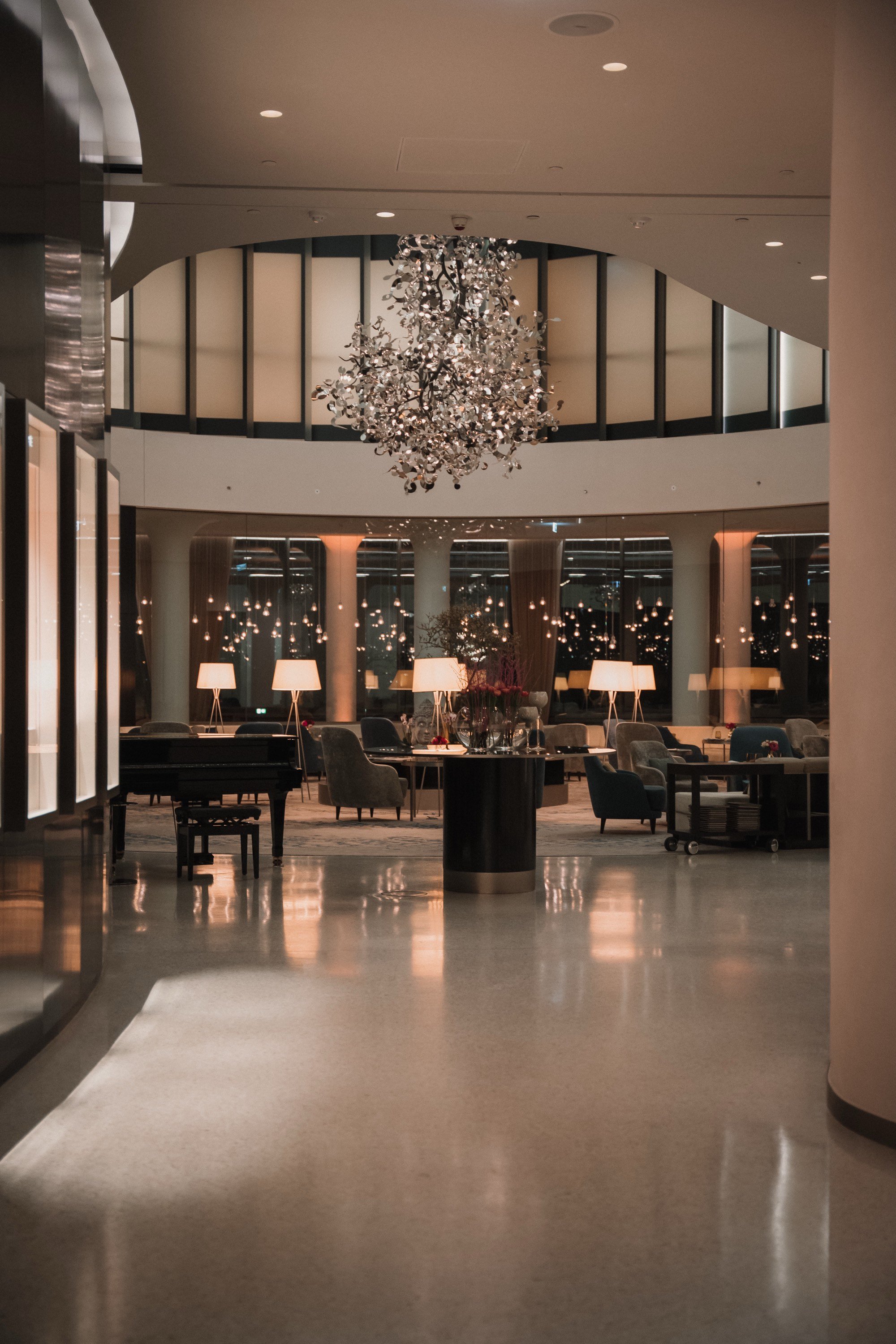 Elegantes Hotel-Lounge mit modernem Kronleuchter, bequemen Sesseln, Stehlampen und reflektierenden Fenstern bei Nacht.