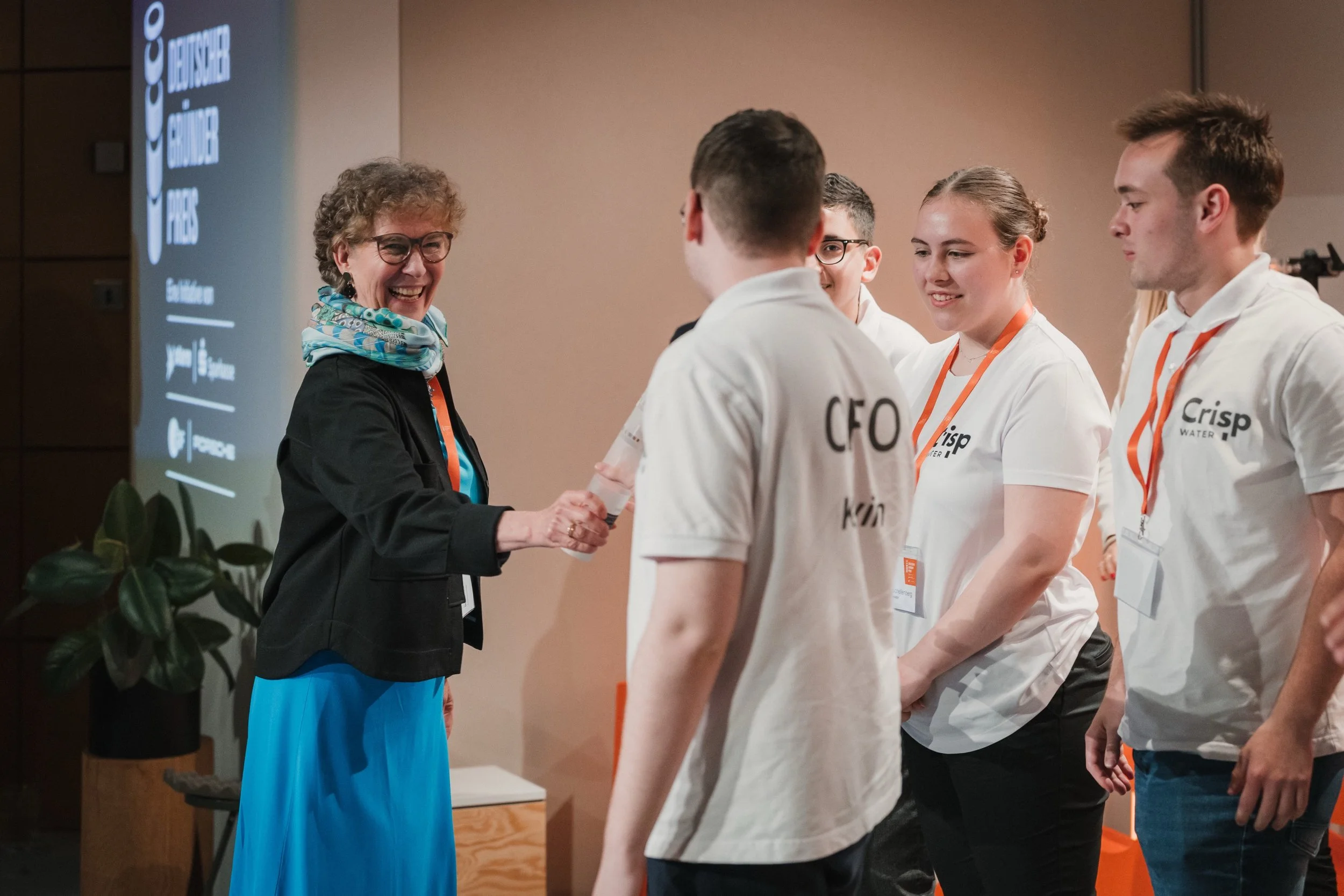 Eine Frau beim Händeschütteln mit einem Jugendlichen auf einer Bühne, umgeben von weiteren Jugendlichen in weißen T-Shirts, bei einer Preisverleihung oder einer Zeremonie im Rahmen eines Events.